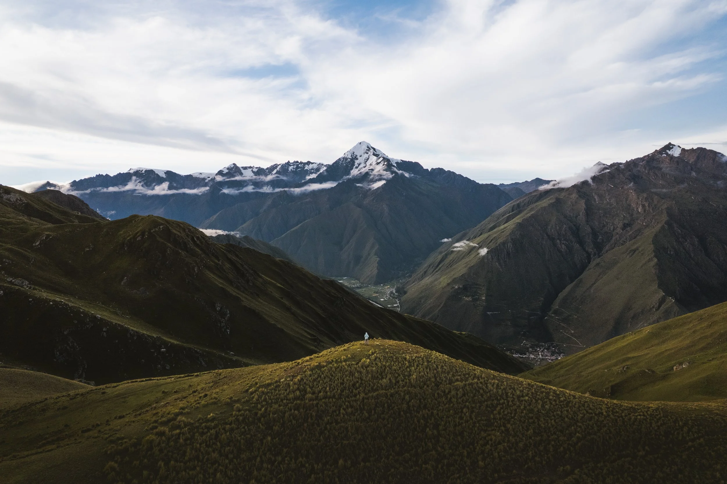 Scenic view of green rolling hills in foreground, vast mountain range with snow-capped peaks, some clouds, and a partly cloudy sky in the background.