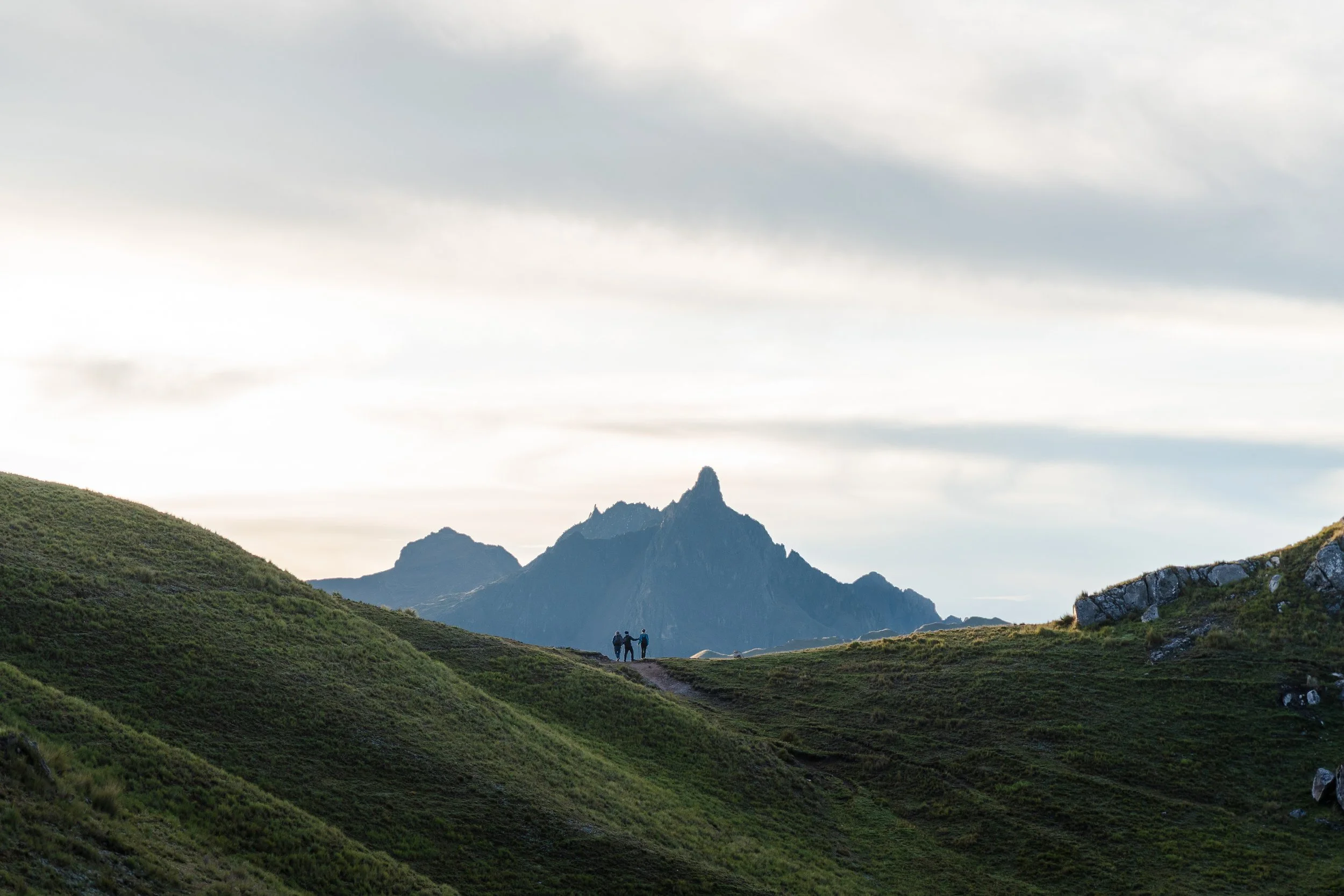 Three hikers stand on the Ancascocha trail in Cusco, Peru on green hills with a mountain range and cloudy sky in the background.