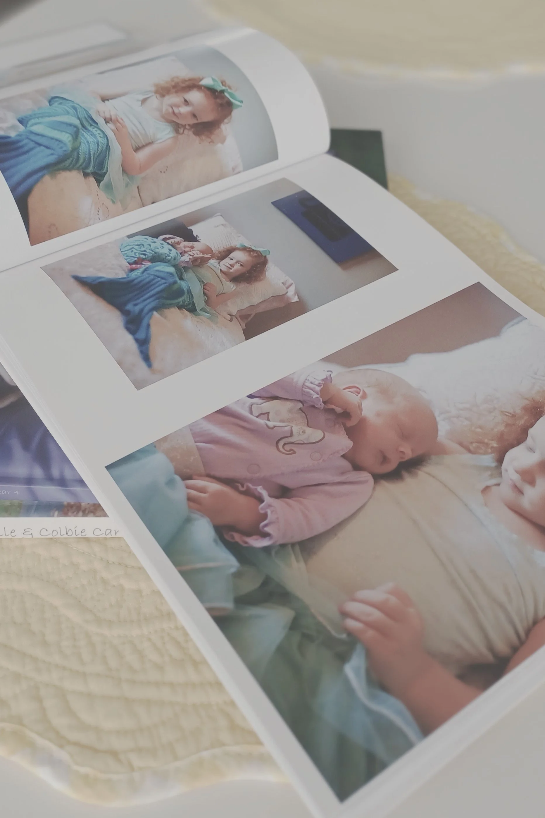 Open photobook on a table displaying two pages of photos. One page shows a young girl with curly hair in a turquoise dress lying on a bed. The other page shows the same girl next to a sleeping baby in a pink outfit.
