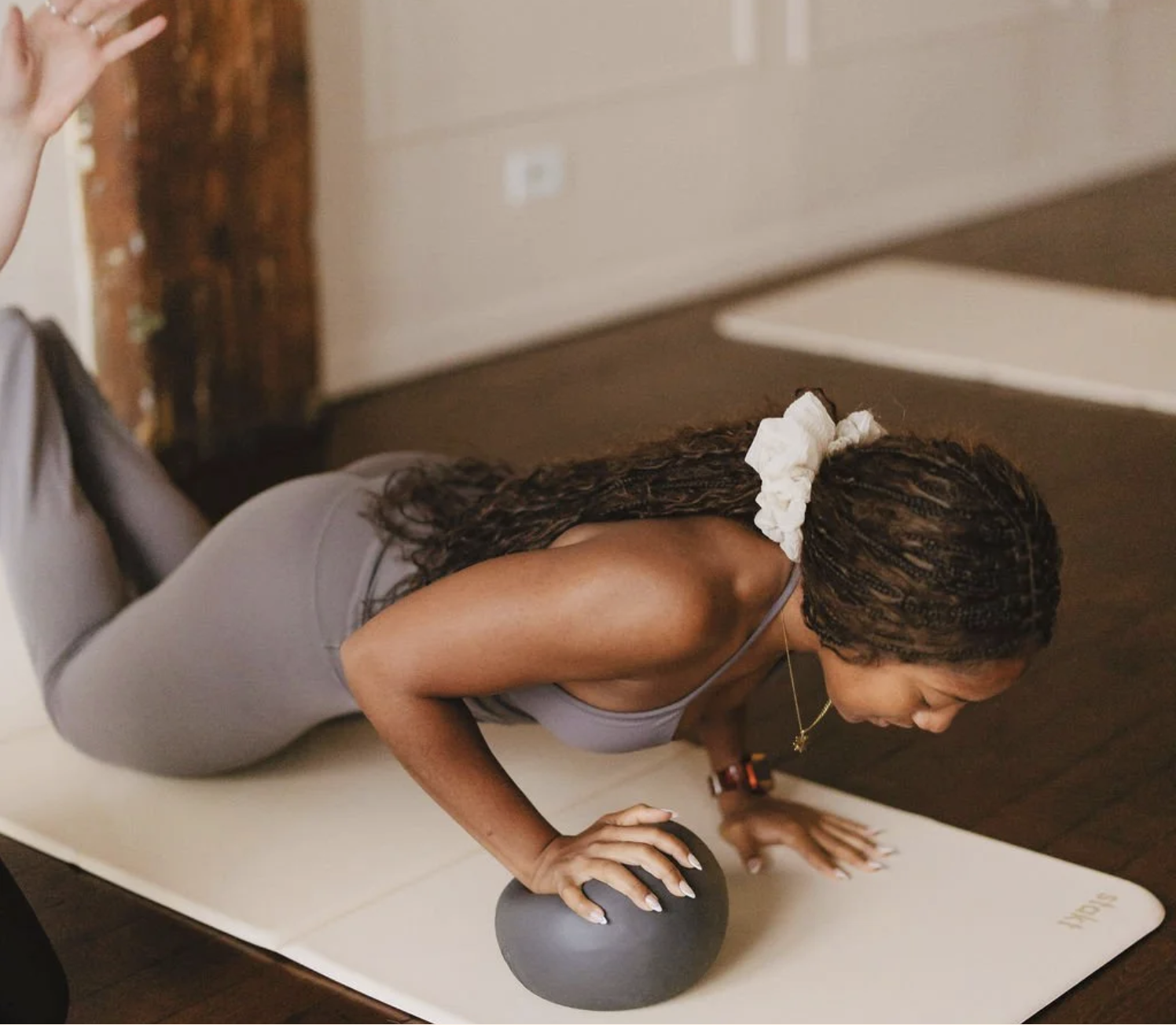 A woman with long braided hair in a ponytail secured with a white scrunchie, wearing a gray tank top and gray leggings, performing a push-up with her hands on an exercise ball on a white yoga mat in a room.