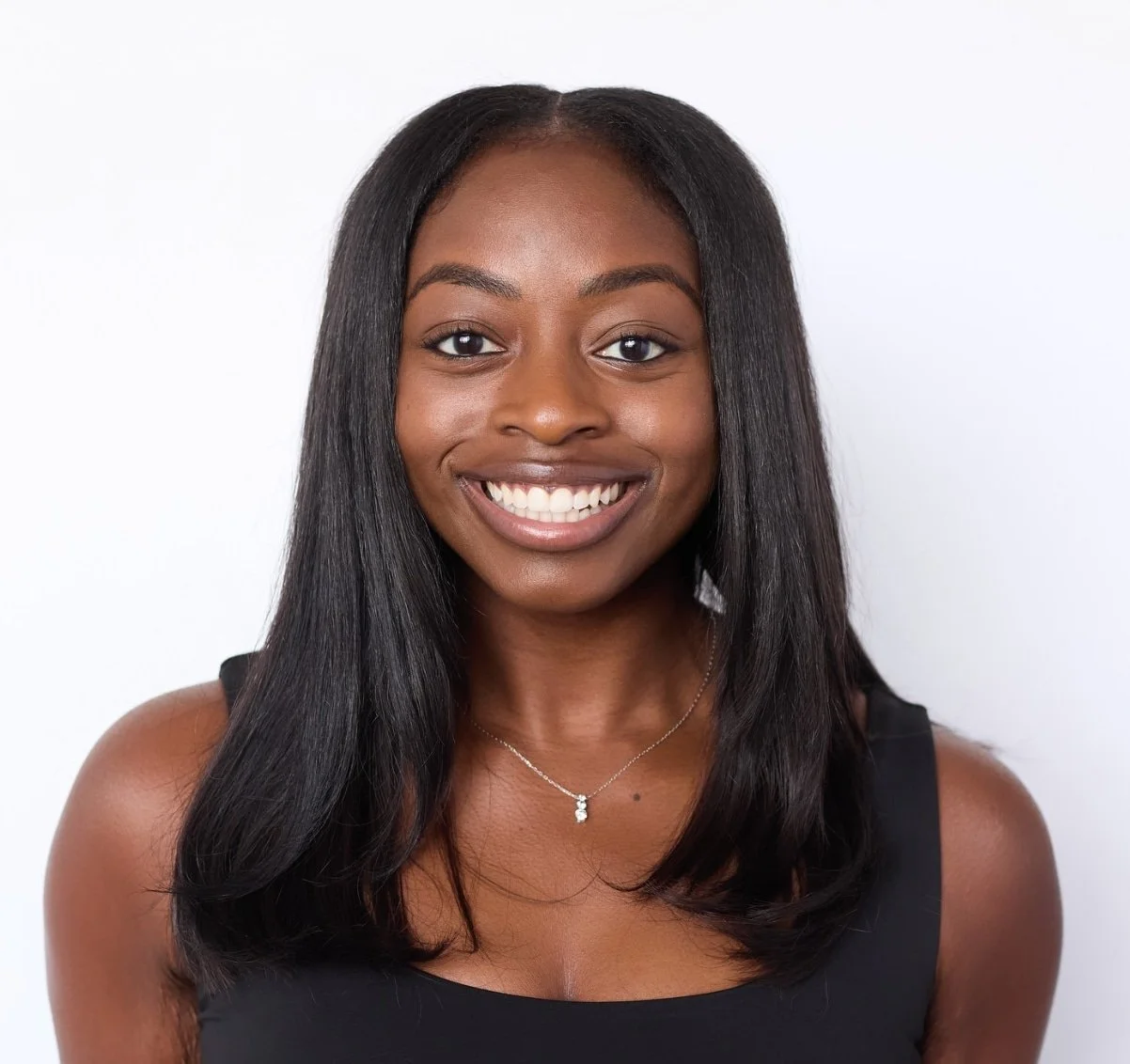 A woman with long black hair wearing a black sleeveless top and a silver necklace smiling against a white background.