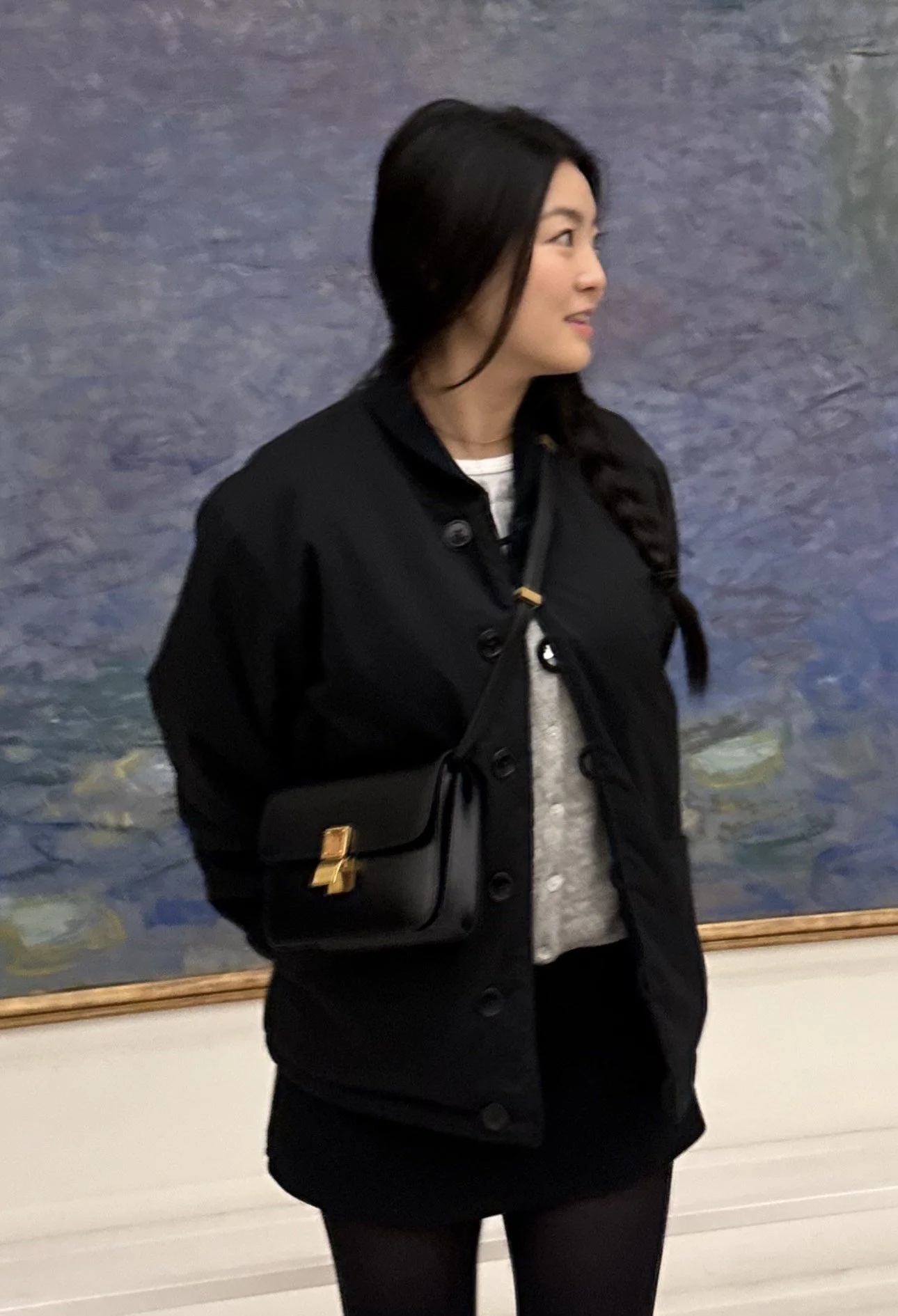 A woman with long dark hair wearing a black coat and carrying a black purse stands in front of a colorful painting.