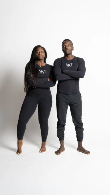 Two individuals standing side by side with arms crossed, wearing black long-sleeve shirts and pants, posing against a white background.