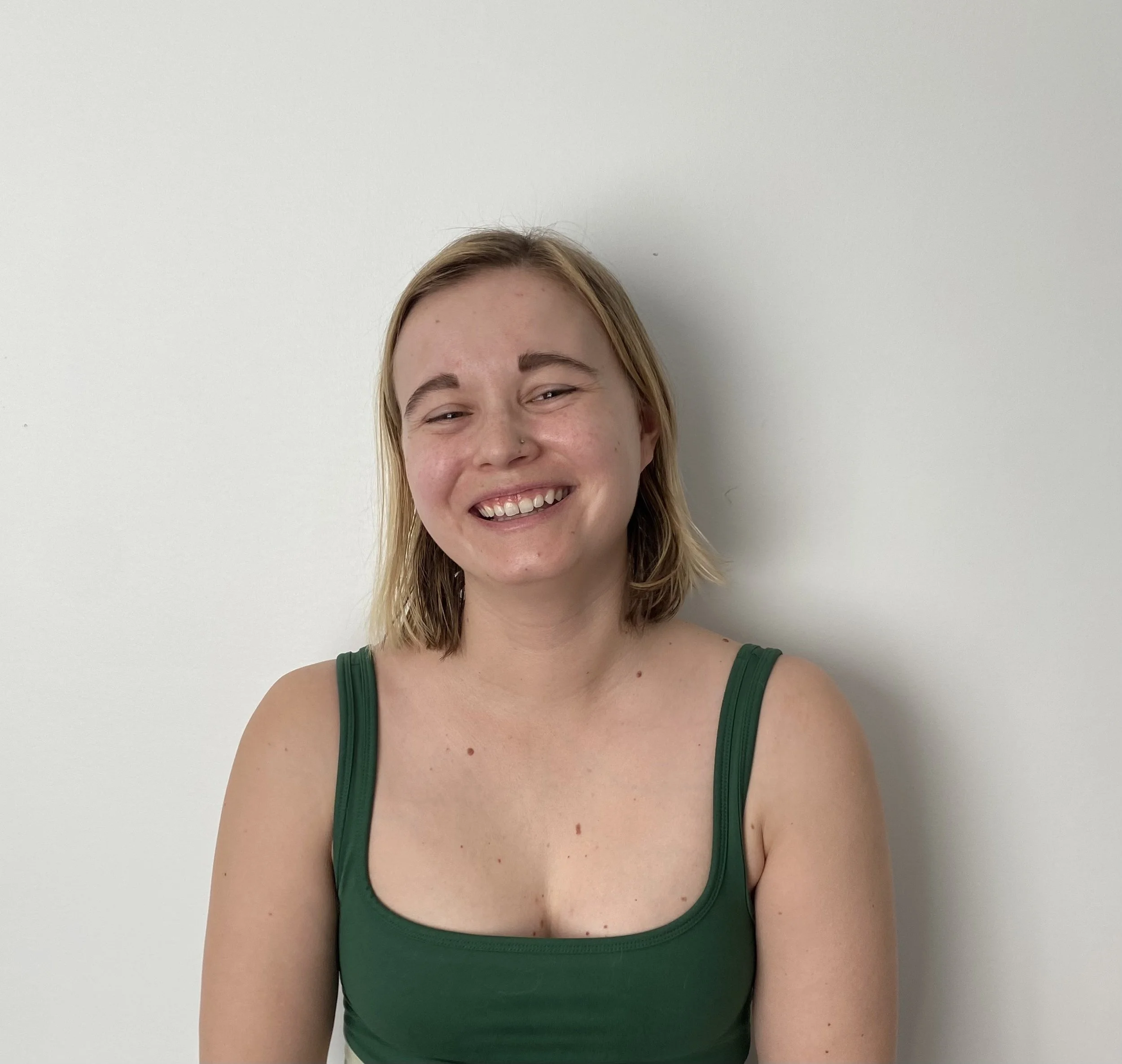 A woman with shoulder-length blonde hair, smiling, wearing a green tank top, standing against a plain white wall.