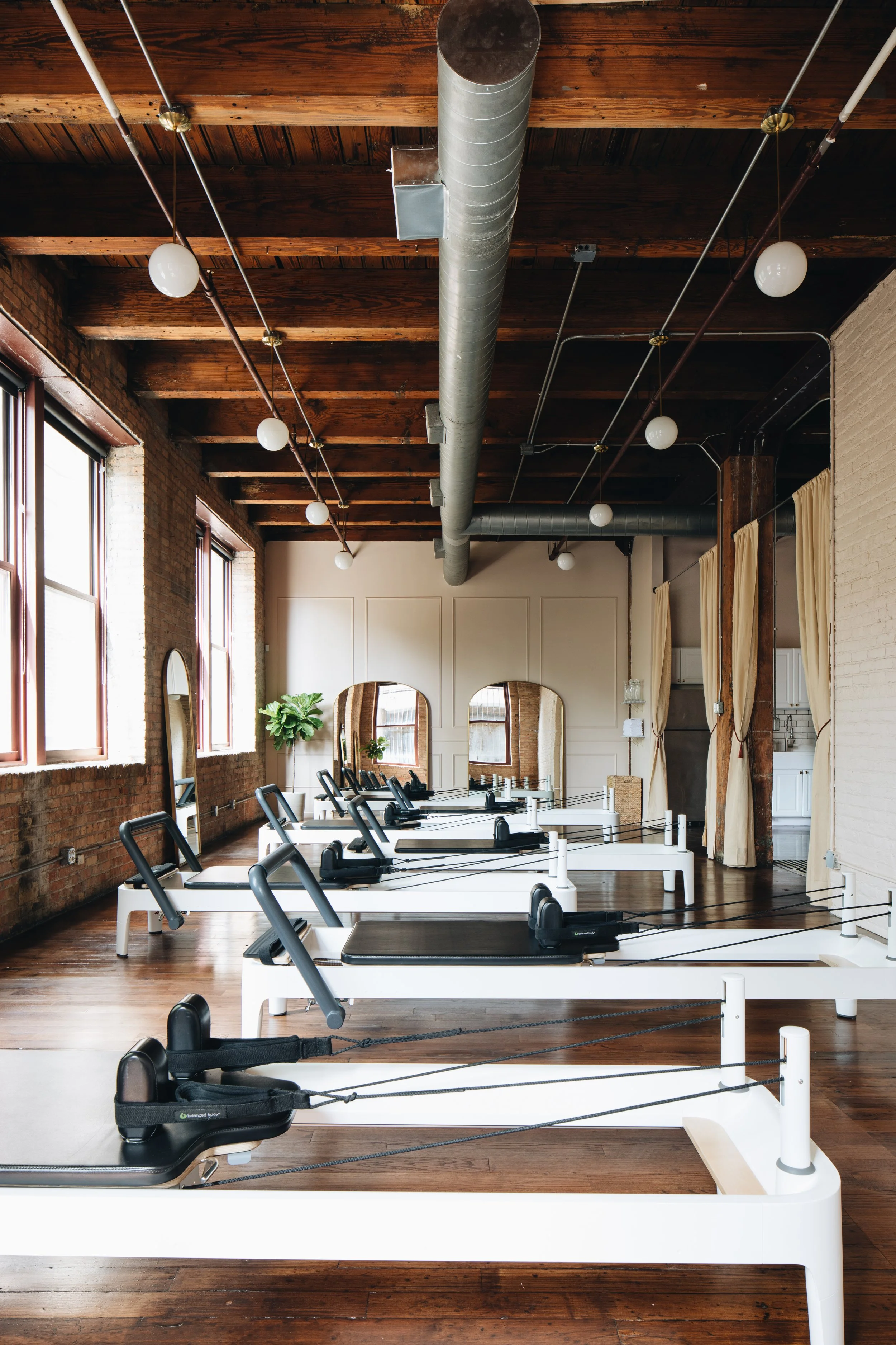 Row of pilates reformer fitness machines with black padding in a bright industrial-style fitness studio with exposed brick walls, large windows, and wood ceiling beams.