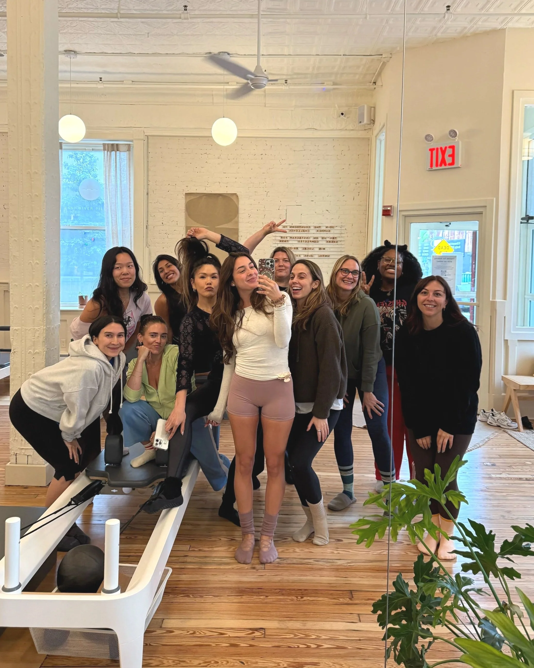 A group of diverse women stand together in a fitness studio, taking a mirror selfie. They are smiling and making playful gestures. The studio has white brick walls, large windows with curtains, a ceiling fan, and wooden floors. A plant is visible in the foreground, and fitness equipment is in the background.