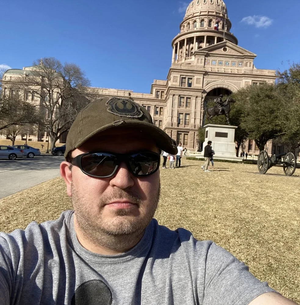 Kevin Golden in a cap and sunglasses taking a selfie in front of the Texas State Capitol in Austin
