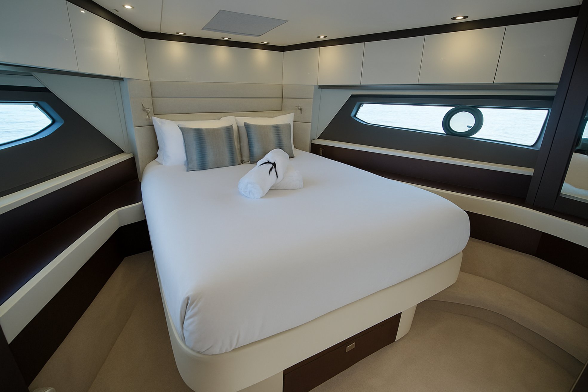 Luxurious bedroom inside a yacht with large windows showing water outside, a neatly made bed with white linens, two rolled towels with a ribbon, and decorative pillows.