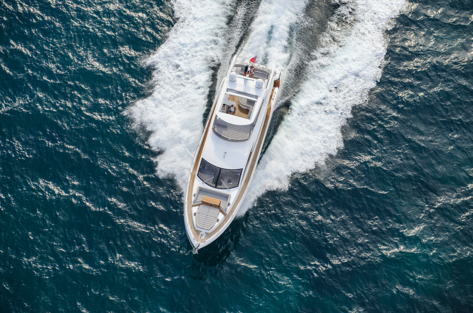 Two people stand on the steps of a luxury yacht in a marina with city skyscrapers in the background, waving and smiling.