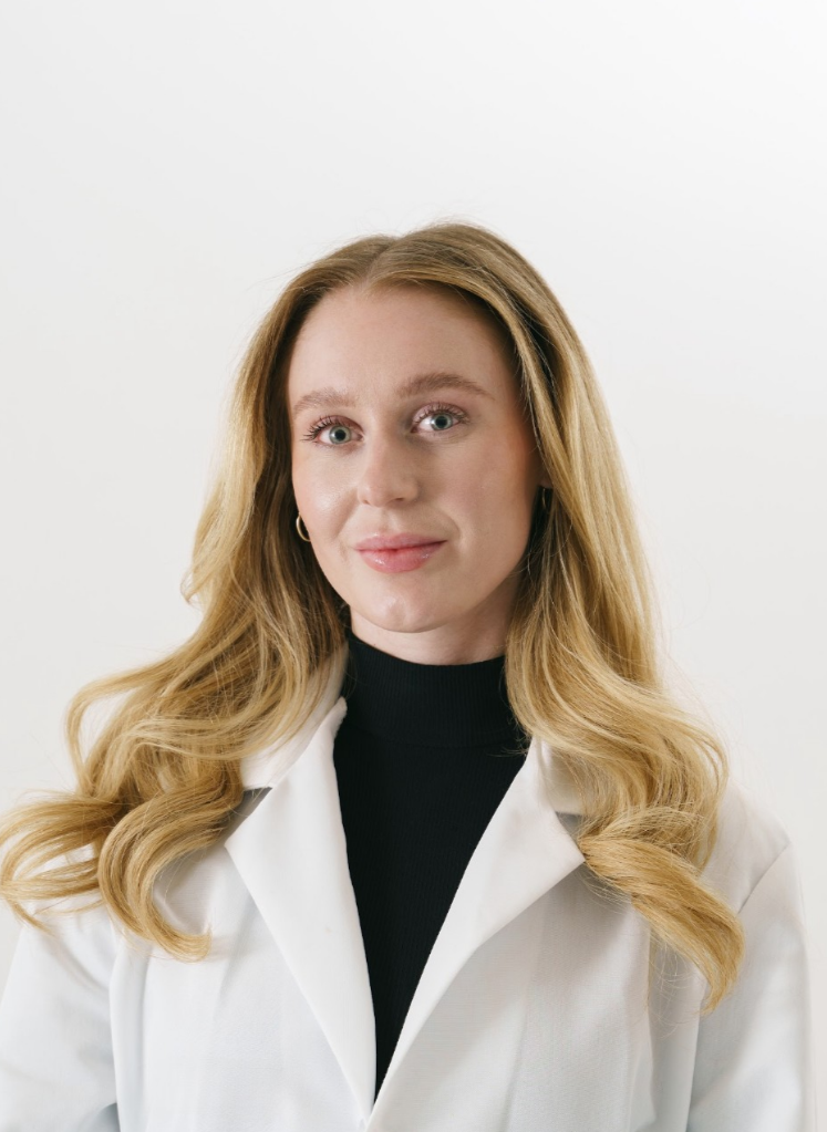 Portrait of a woman with long wavy blonde hair, wearing a white lab coat over a black turtleneck, standing against a plain white background.