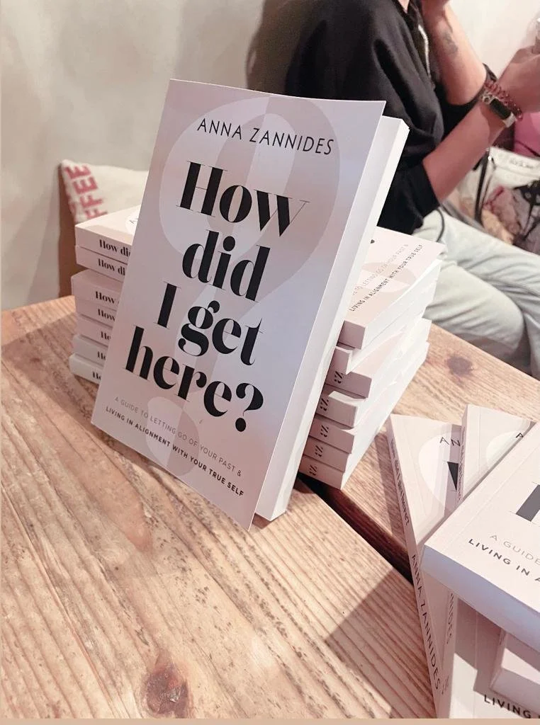 Stack of books titled 'How Did I Get Here?' by Anna Zannides on a wooden table in a cafe setting.