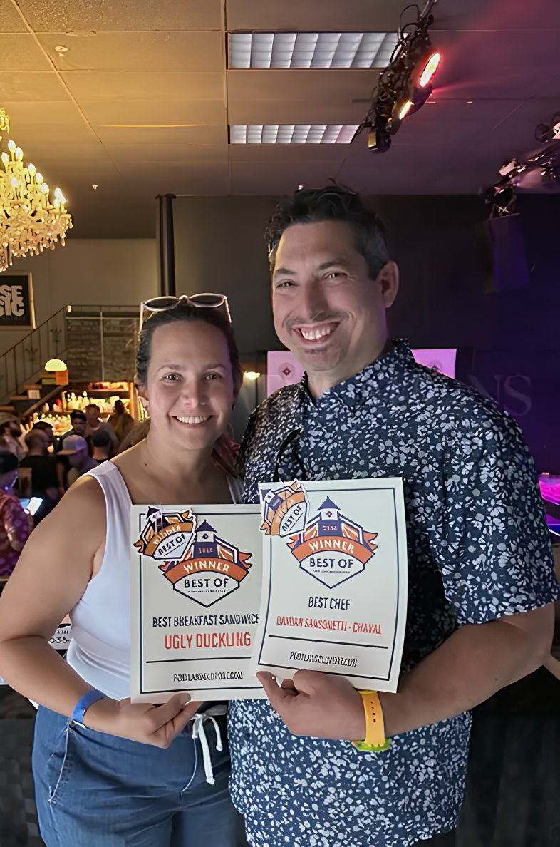 Two smiling people holding award certificates at an event, with a woman on the left and a man on the right, in a lively indoor setting with a bar and group of people in the background.