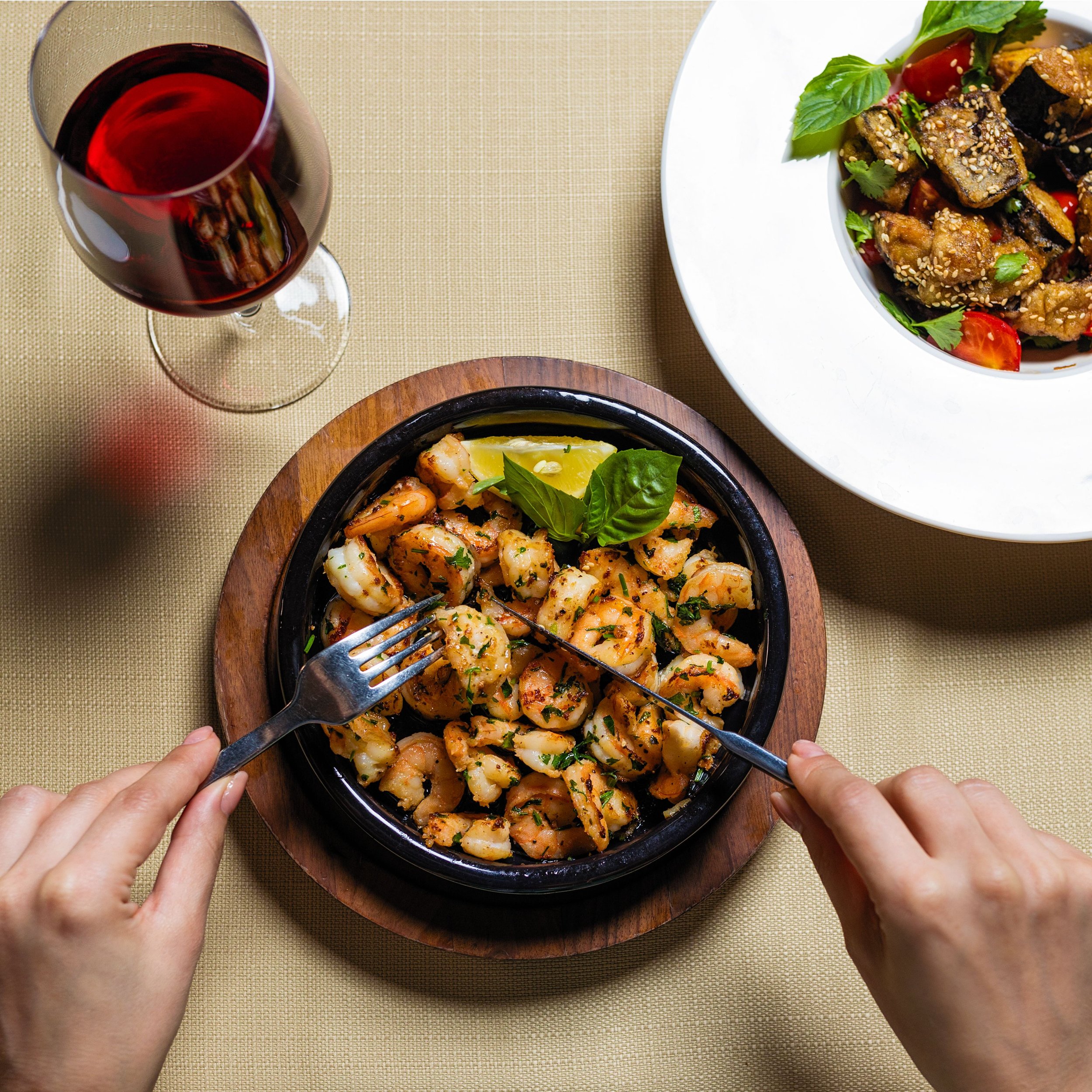 Hands holding a fork and spoon over a bowl of shrimp dish on a round wooden tray, with a glass of red wine nearby and a white bowl of vegetable salad with cherry tomatoes and basil leaves on a beige tablecloth.