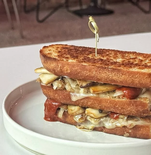 A sandwich made with toasted bread and melted cheese, with visible vegetables and possibly meat, secured with a decorative toothpick, served on a white plate.