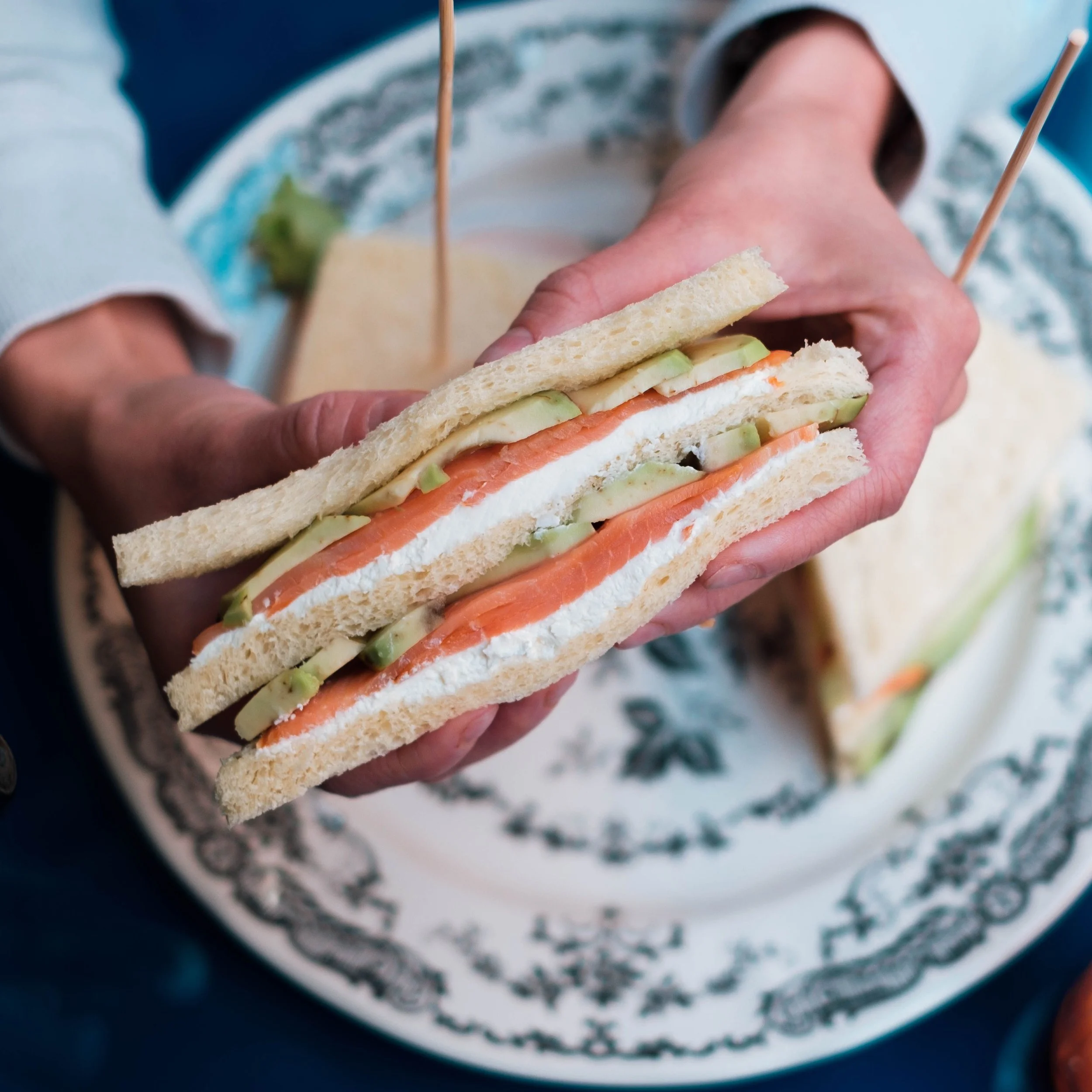 Close-up of a person holding a sandwich with slices of cucumber, smoked salmon, and cream cheese on white bread.
