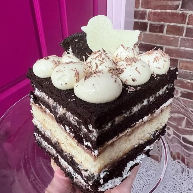 Three-layer slice of black and white cake with white cream and chocolate topping, decorated with a white duck-shaped topper and cookie decorations, on a glass cake stand.