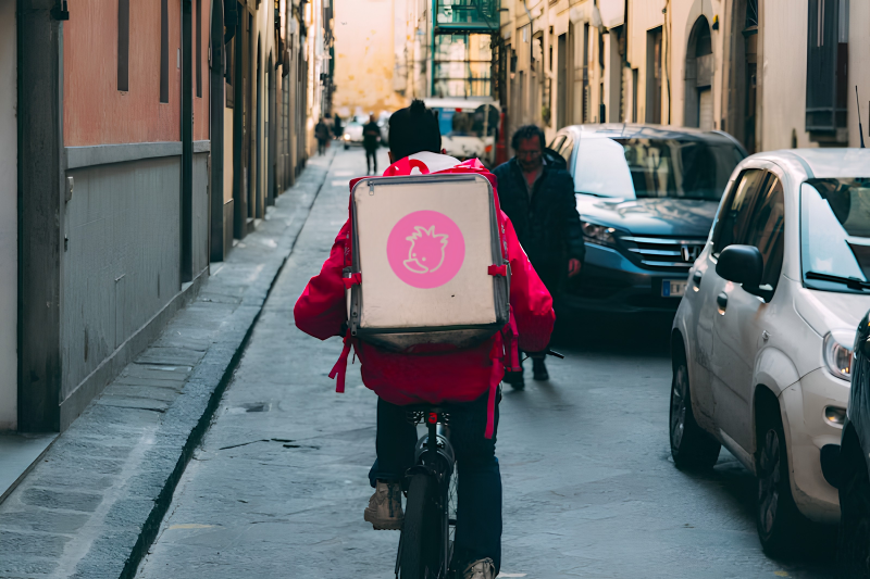 Person riding a bicycle on a narrow city street with parked cars and pedestrians, carrying a backpack with a logo of a pink hedgehog.
