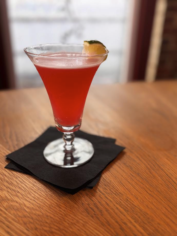 Red cocktail with lime wedge garnish on a black napkin, placed on a wooden table.