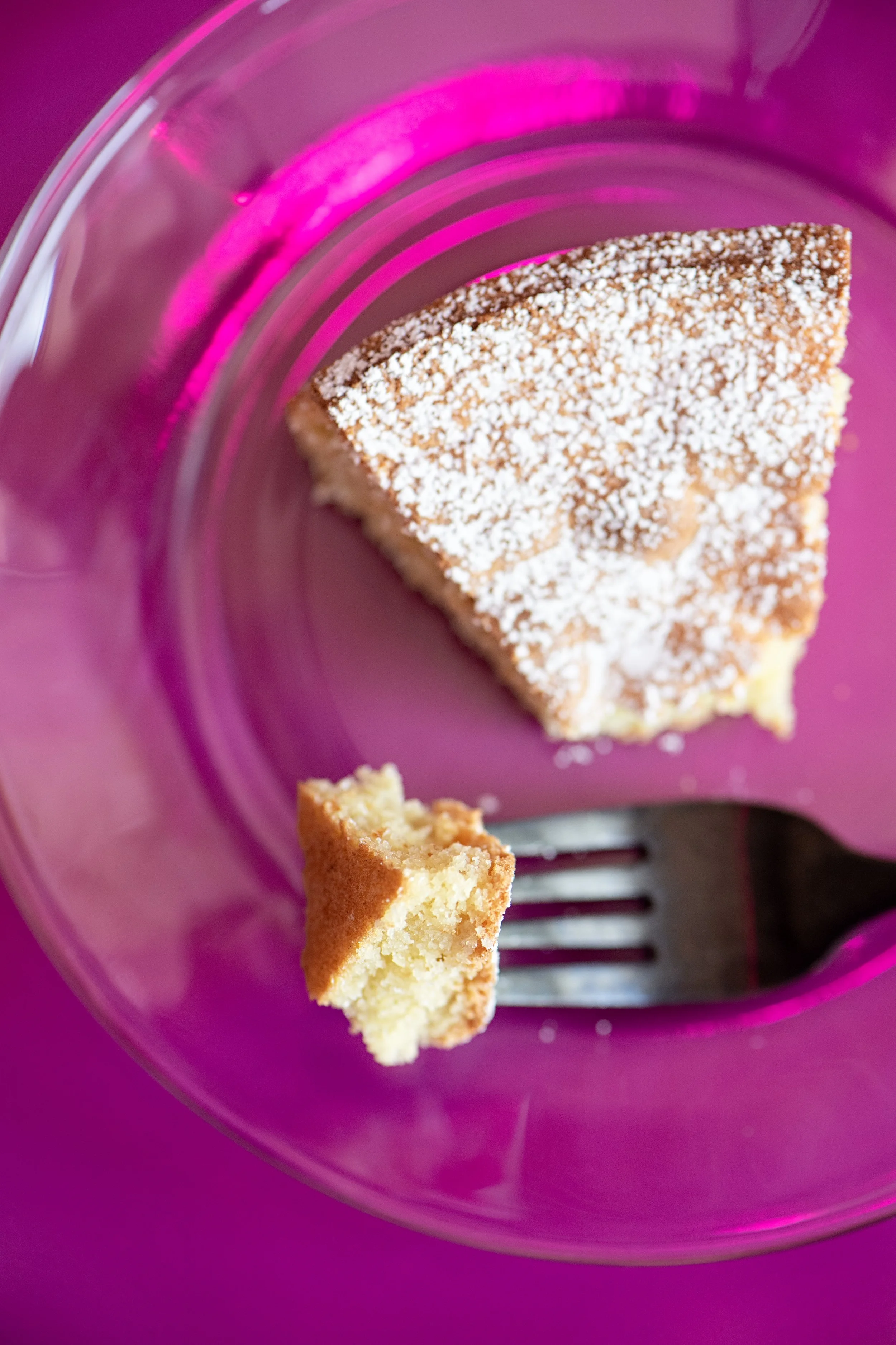 Close-up of a piece of vanilla cake with white frosting crumbs on top, placed on a purple plate with a black fork. Part of the cake and a bitten piece are visible.
