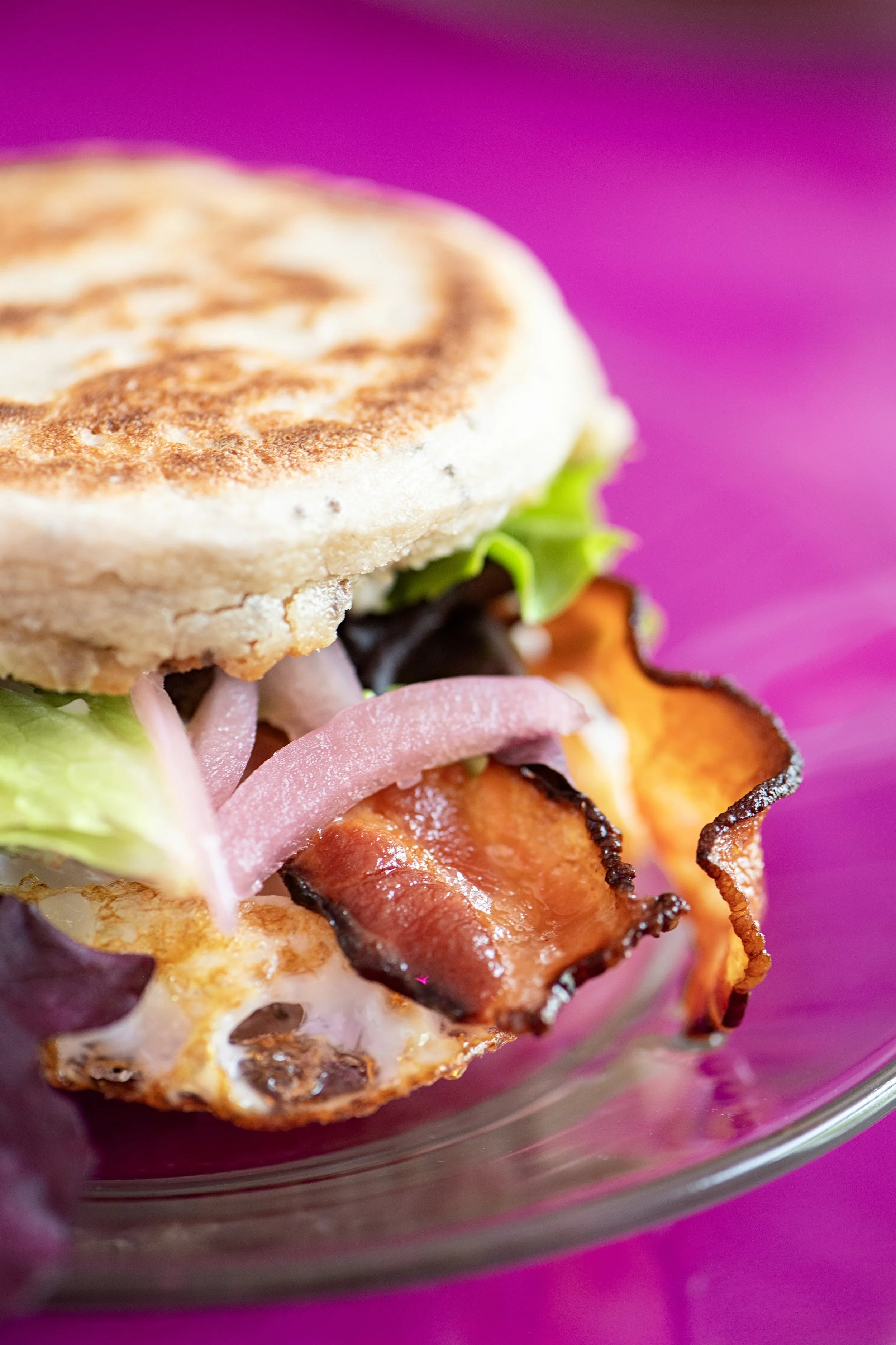Close-up of a breakfast sandwich with fried egg, bacon, lettuce, tomato, and sliced onions on an English muffin, on a purple plate.