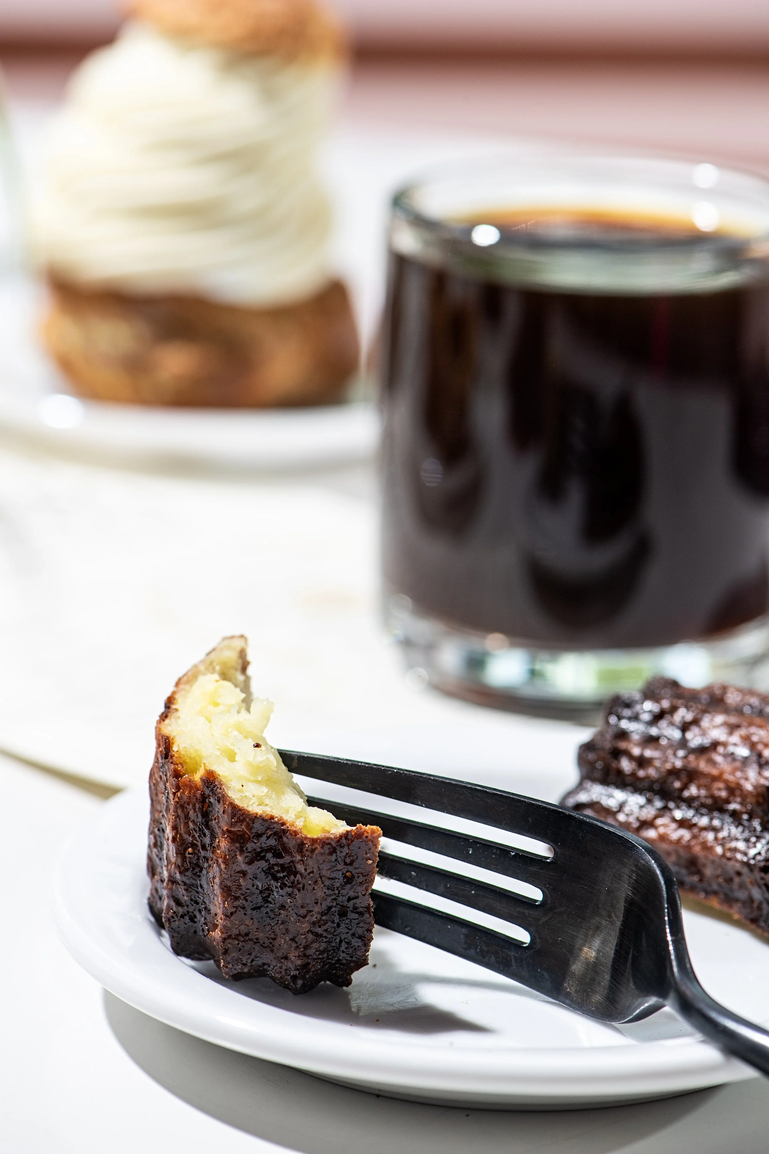Close-up of a piece of shortbread on a black fork, with a white plate underneath, and a blurred background including a glass of dark soda, a stack of cookies, and a glazed meat dish.