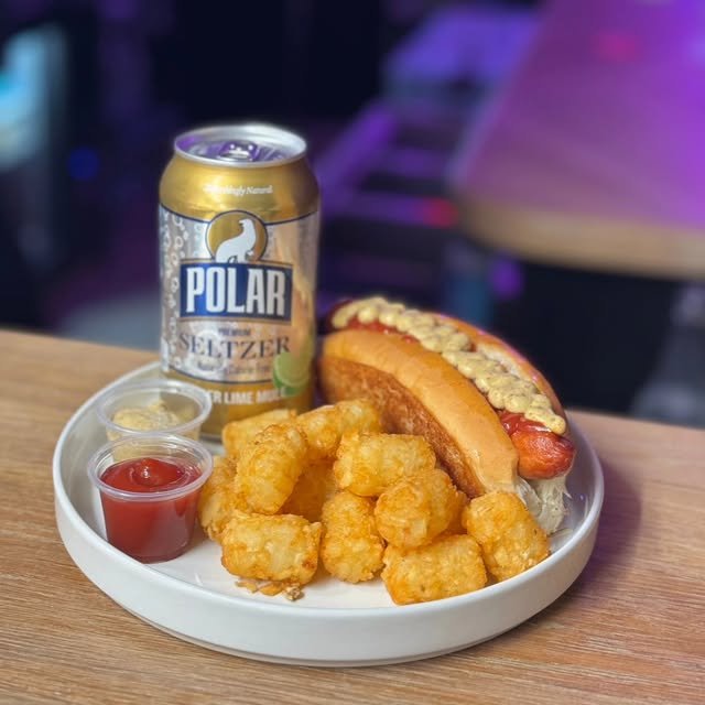 Hot dog with mustard, crispy tater tots with ketchup and a can of Polar seltzer water with lime flavor on a white plate.