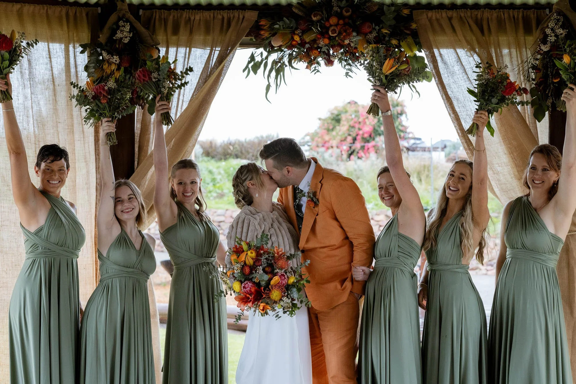 Bride and groom kiss with bridesmaids standing on each side with flowers held in the air