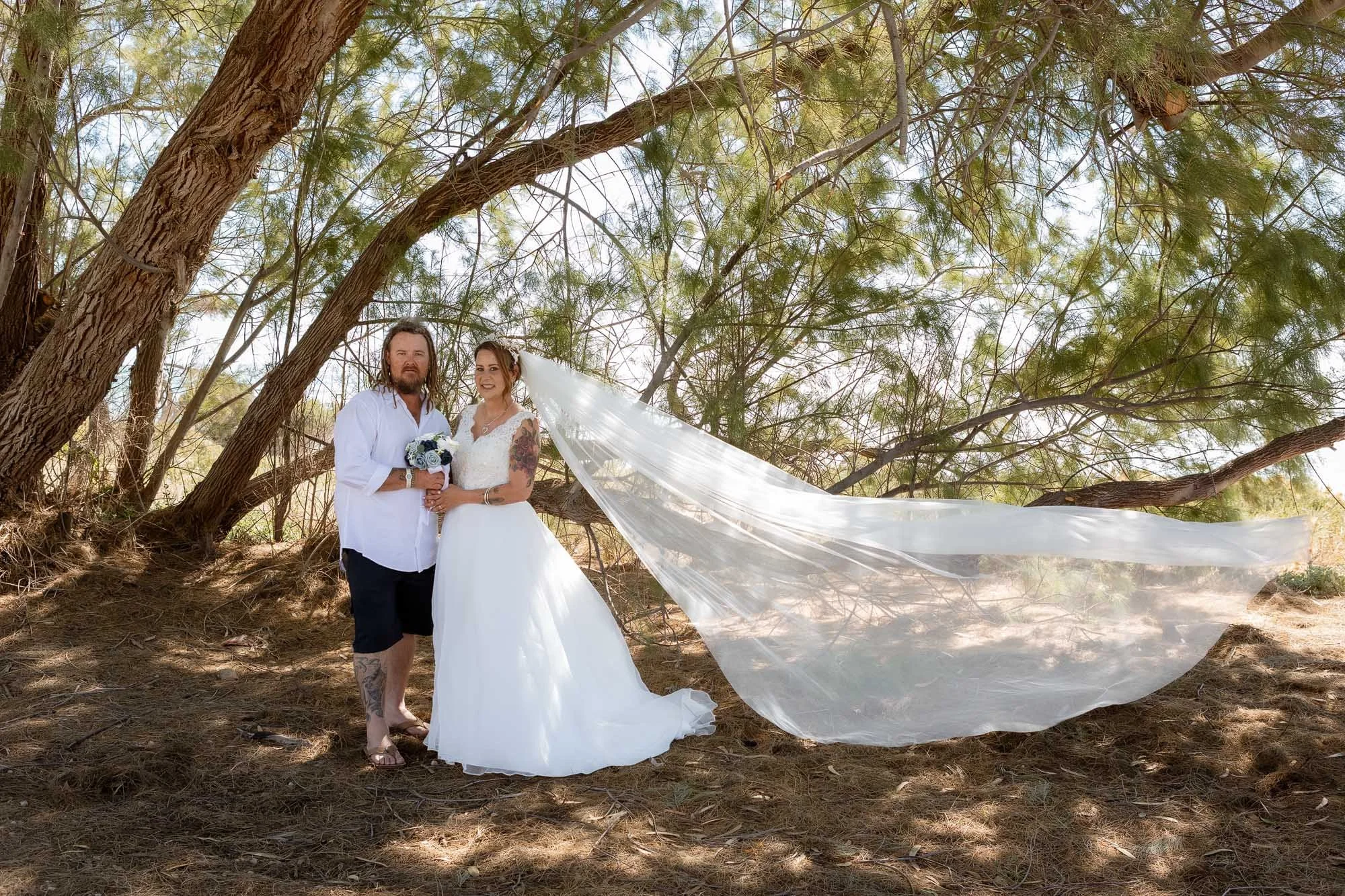 Bride and Groom standing under trees holding flowers with the veil blowing in the wind