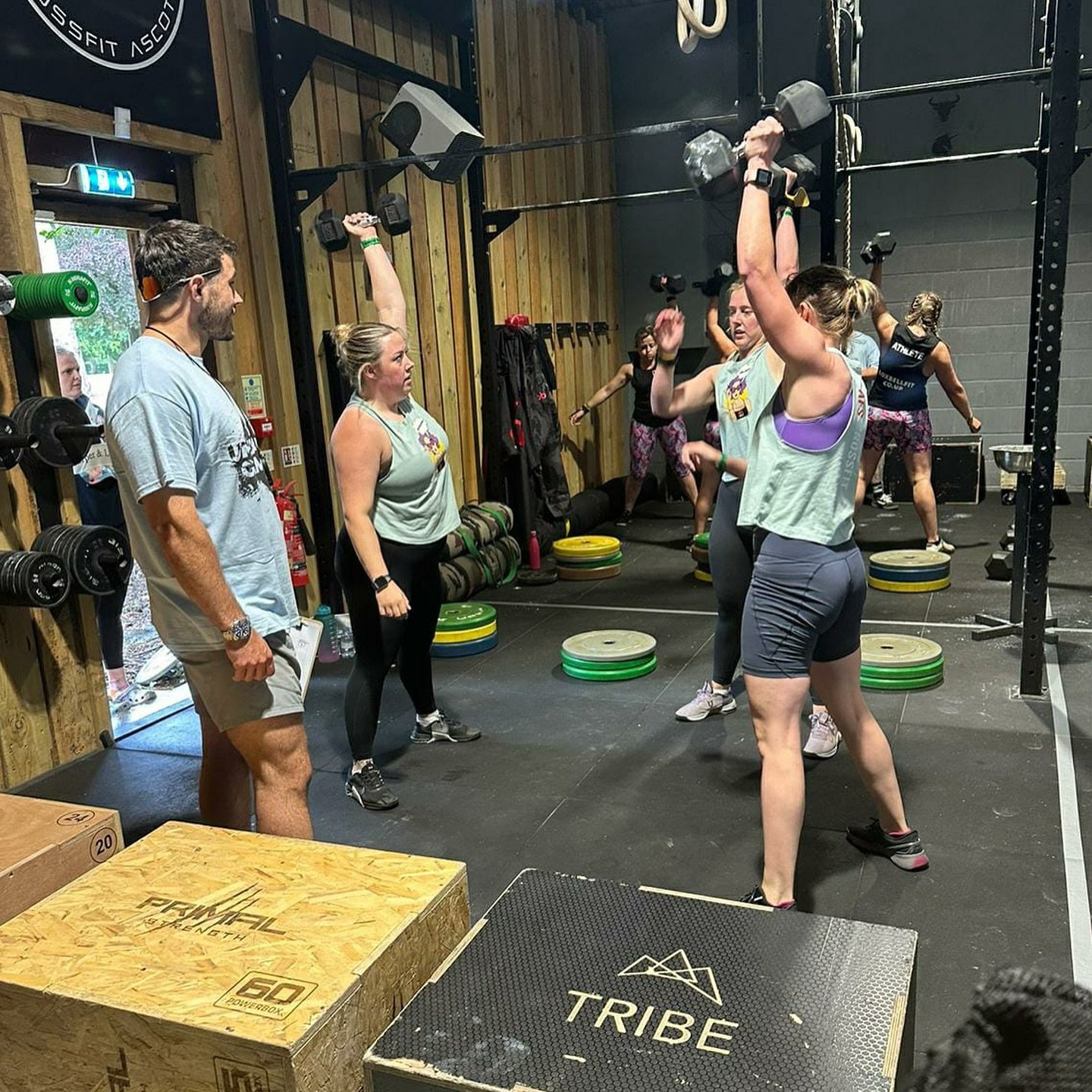 Women participating in a CrossFit workout, lifting dumbbells, in a gym with others in the background, surrounded by weight plates and exercise equipment.