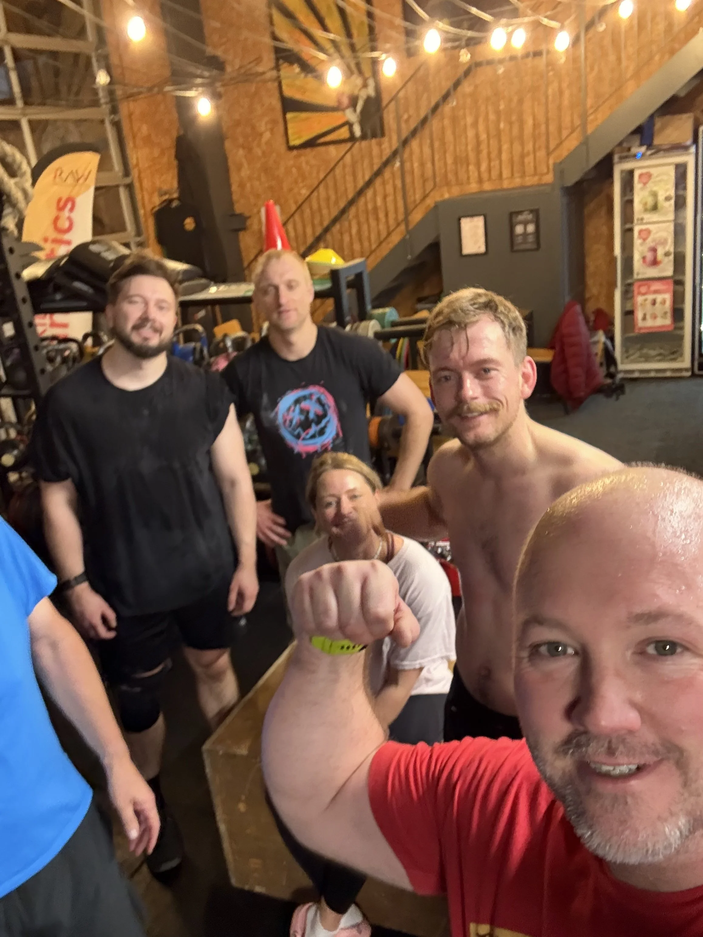 Group of smiling men celebrating post workout selfie, with some flexing their muscles and a man in the foreground making a fist. The background features exposed brick walls, artwork, string lights, and gym equipment.