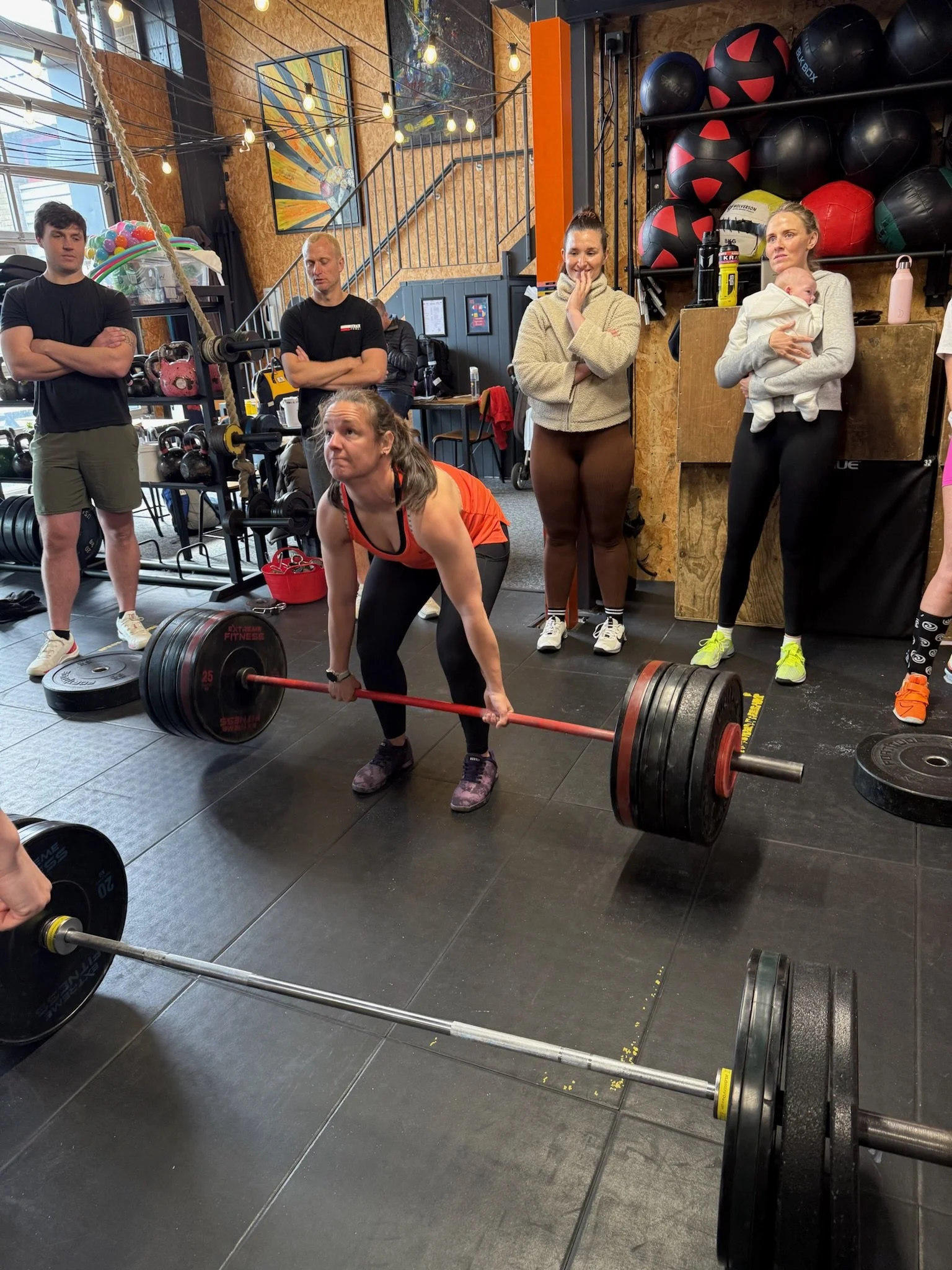 A woman performing a deadlift with a loaded barbell in a gym, surrounded by spectators and gym equipment.