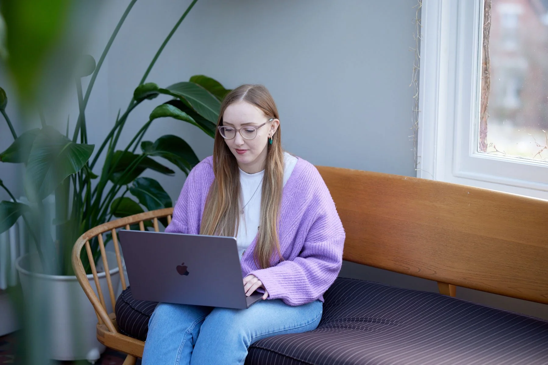 A woman wearing glasses sits with a macbook on her knee looking at the screen