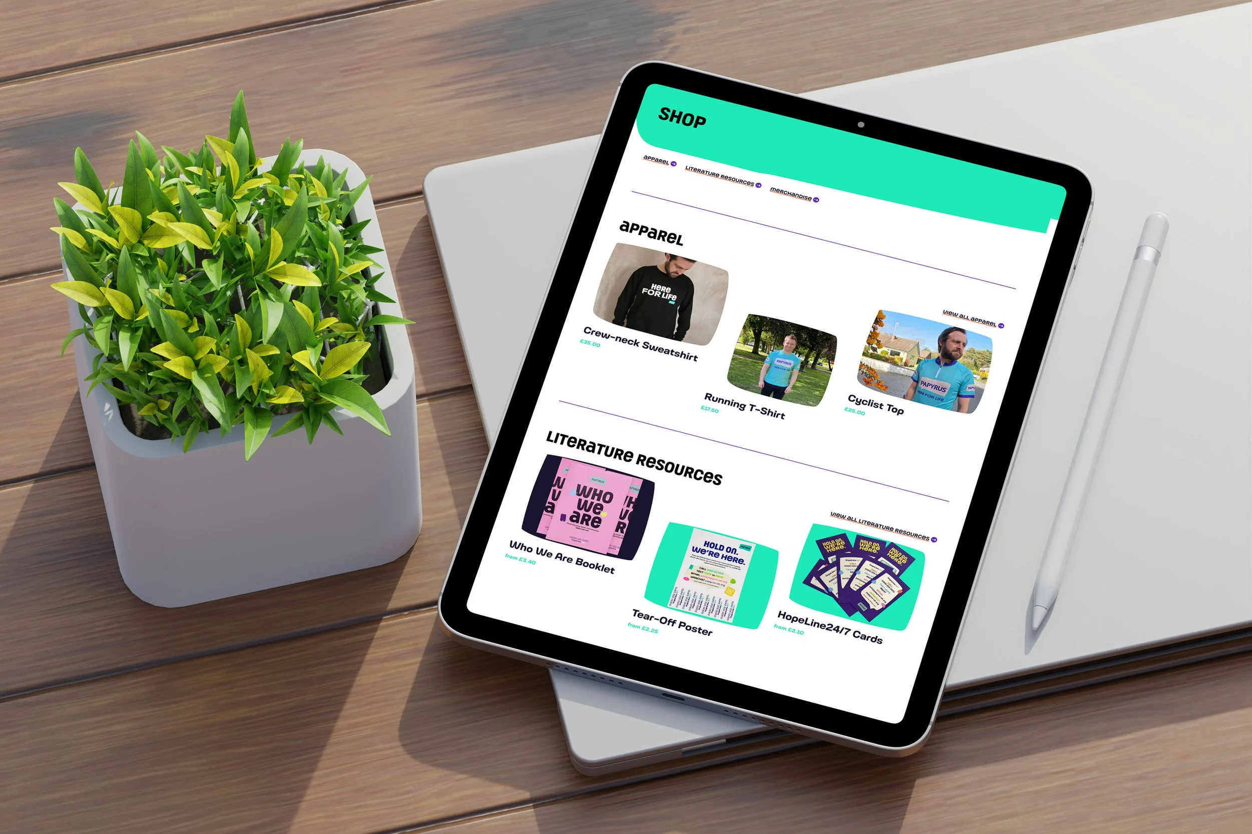 An ipad on top of a closed laptop on a desk next to a stylus and a plant shows a Shop page design from the PAPYRUS website