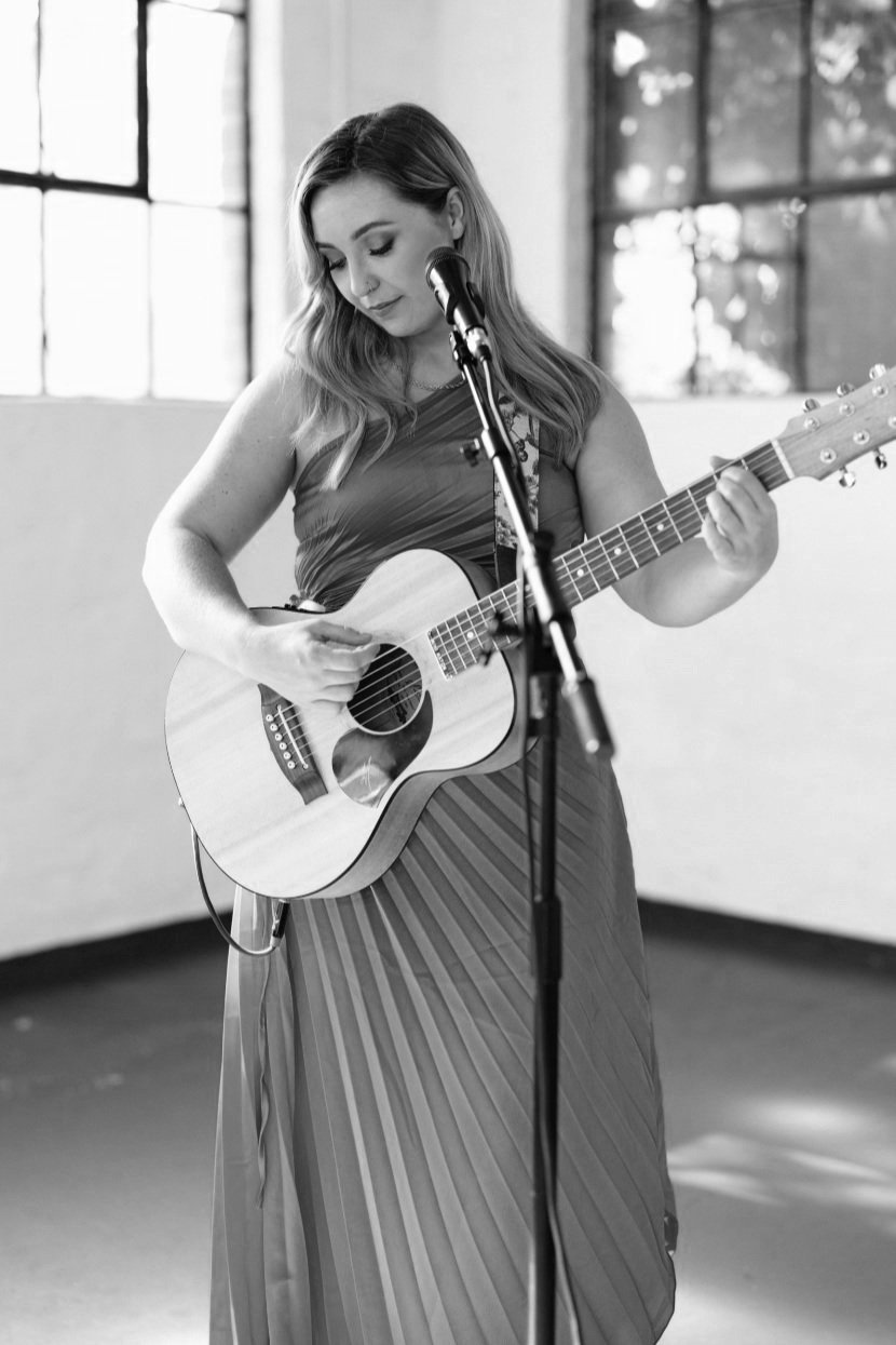 Ruby Miguel with long hair playing an acoustic guitar in front of a microphone in a room with large windows at City Farm in Perth