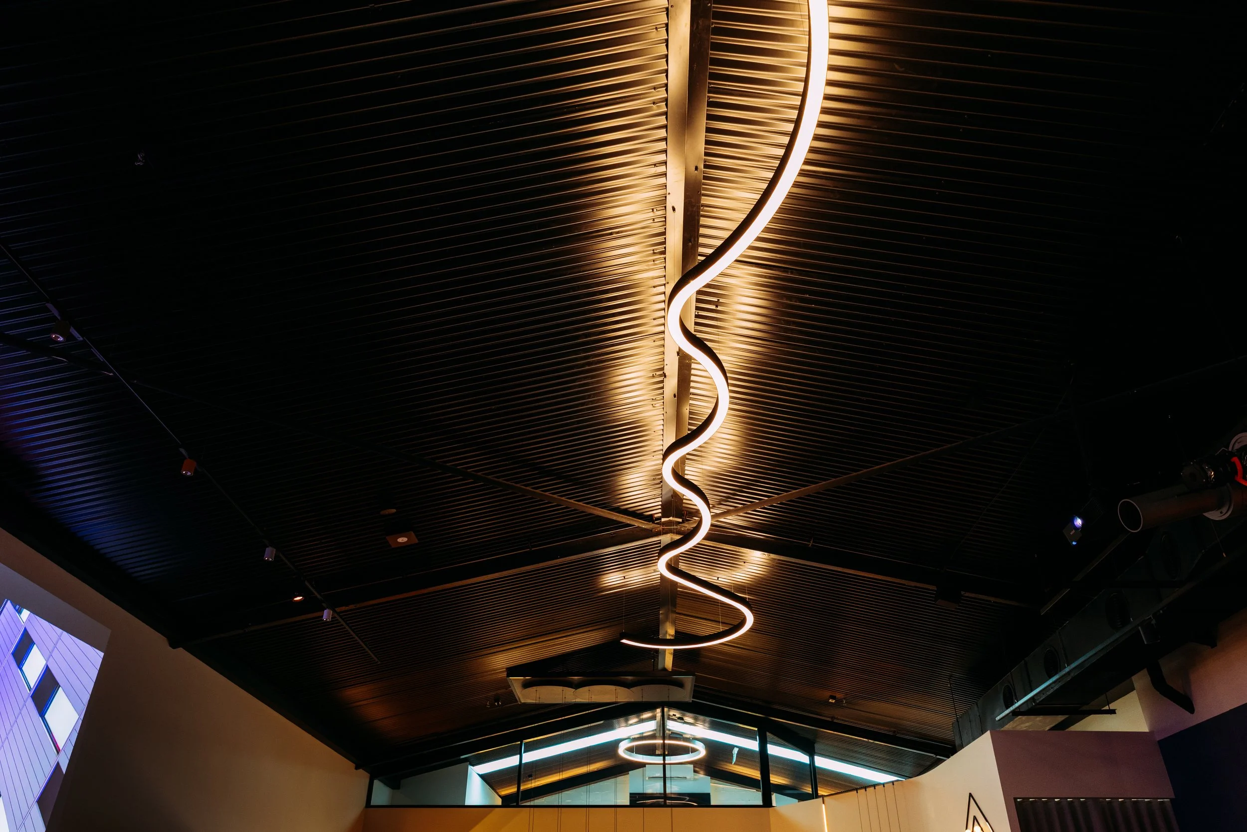 Modern ceiling lighting fixture with wavy neon strip hanging from dark ribbed ceiling in contemporary interior, designed by Raw Process Design, Canberra residential and commercial interior architecture