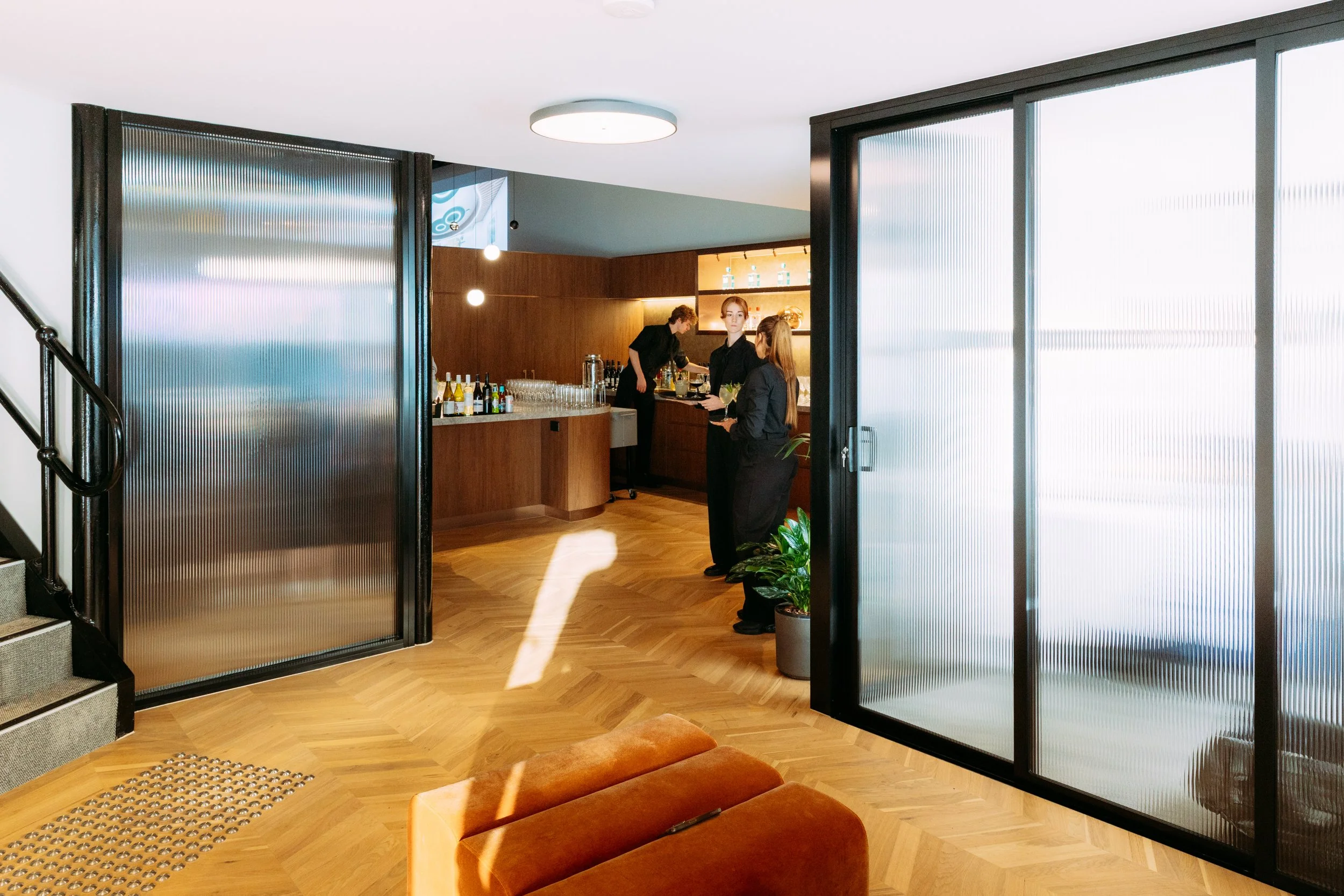 Bar area with three staff preparing drinks, wooden flooring, glass and metal sliding doors, orange armchair, potted plants, illuminated shelves, and staircase on left, designed by Raw Process Design, Canberra interior and architecture