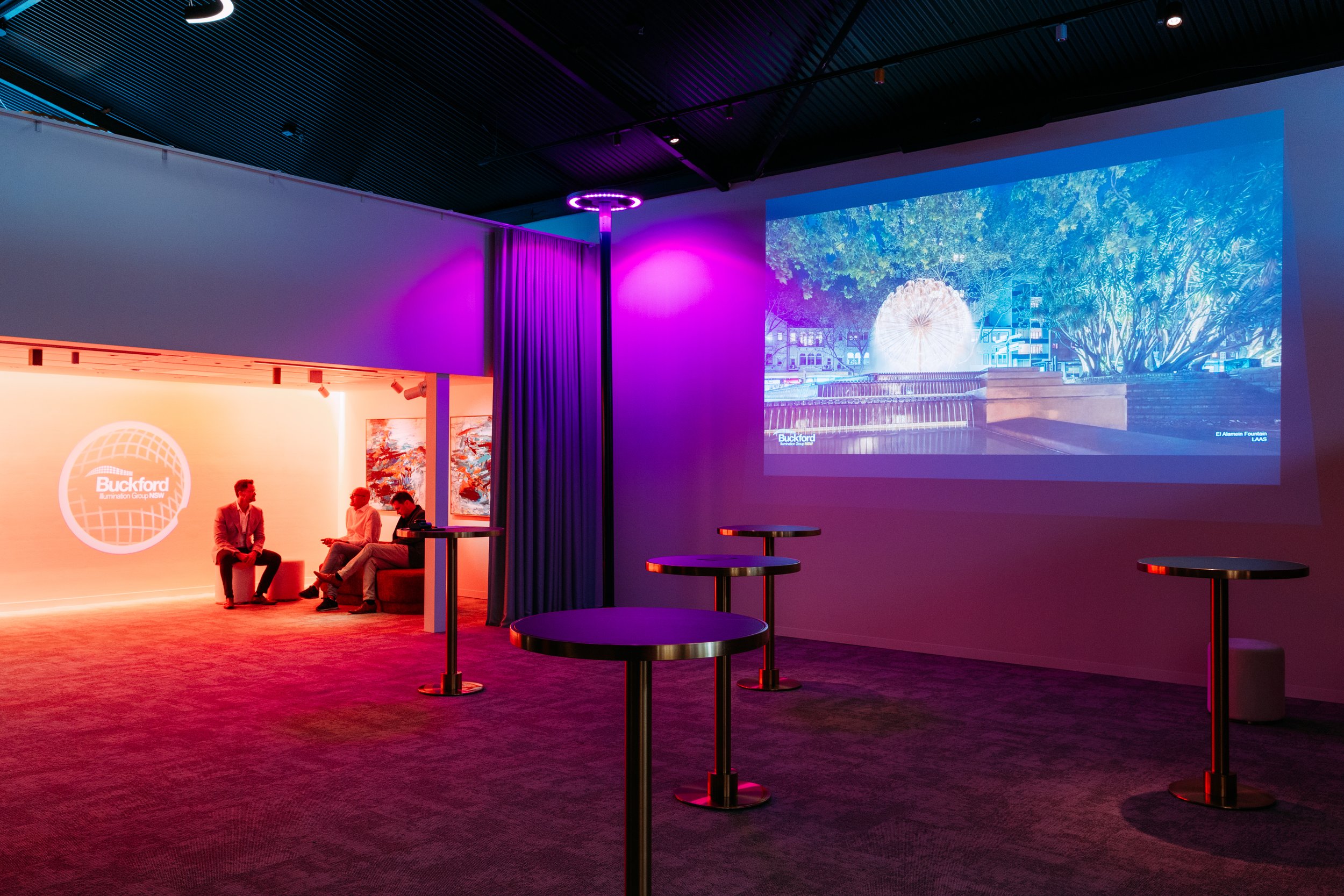 Modern indoor event space with colorful lighting, three people seated on a couch, artwork on the wall, large screen showing nighttime outdoor scene with dandelion sculpture and trees, high tables scattered, designed by Raw Process Design, Canberra