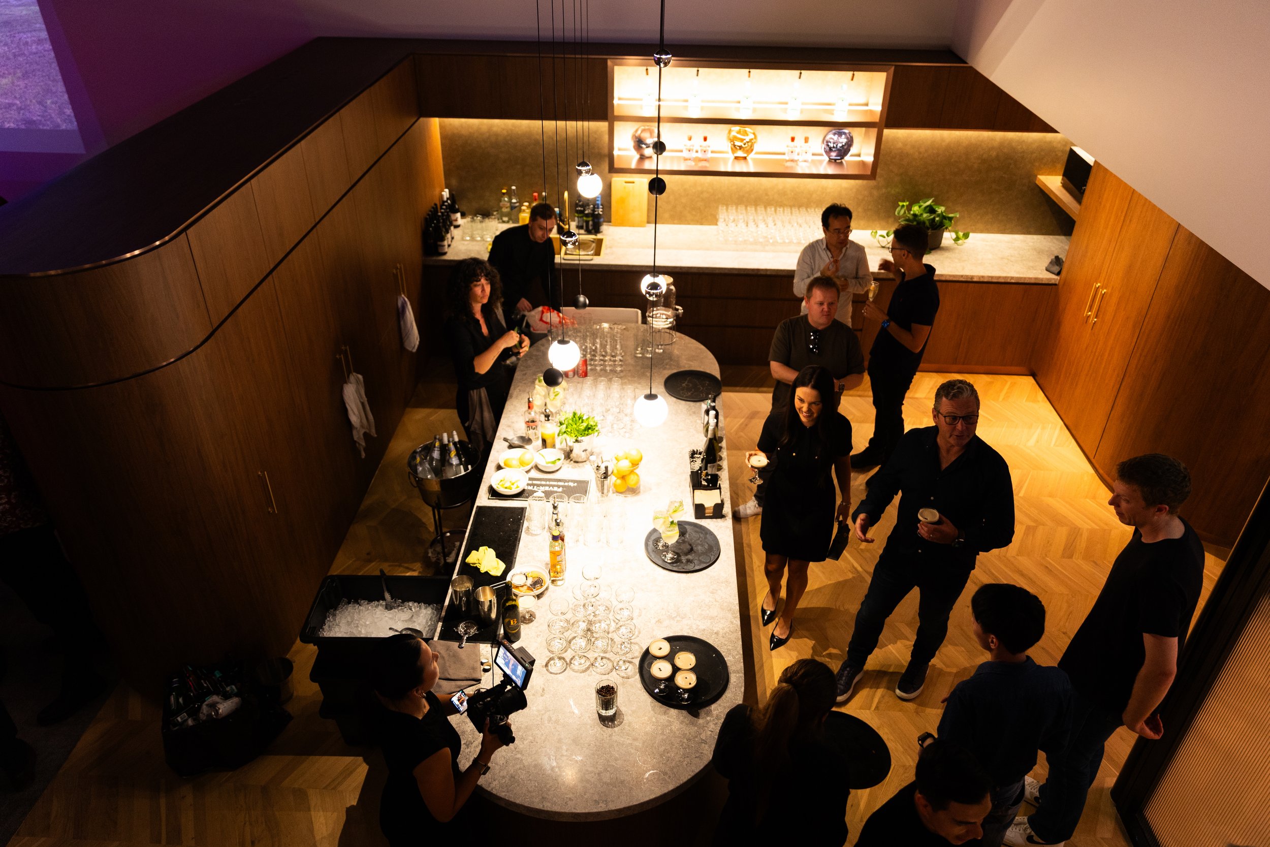 People socializing in a modern home bar with large marble counter, wooden cabinets, pendant lights, and display shelves with glassware, designed by Raw Process Design, Canberra residential interior and architecture