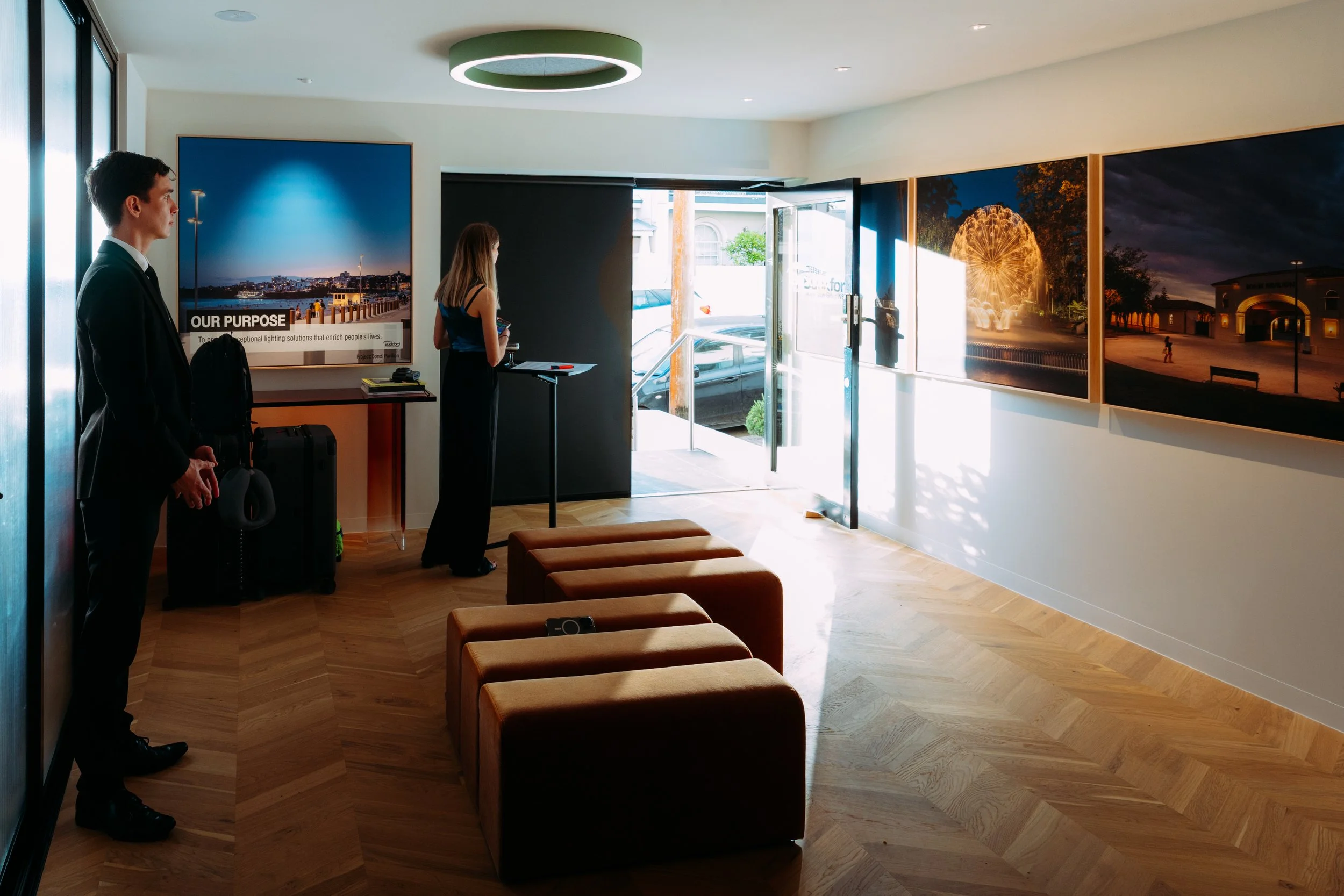 Interior of art gallery with framed photographs on walls, doorway leading outside, two people waiting, and sunlight streaming in, designed by Raw Process Design, Canberra residential and interior architecture