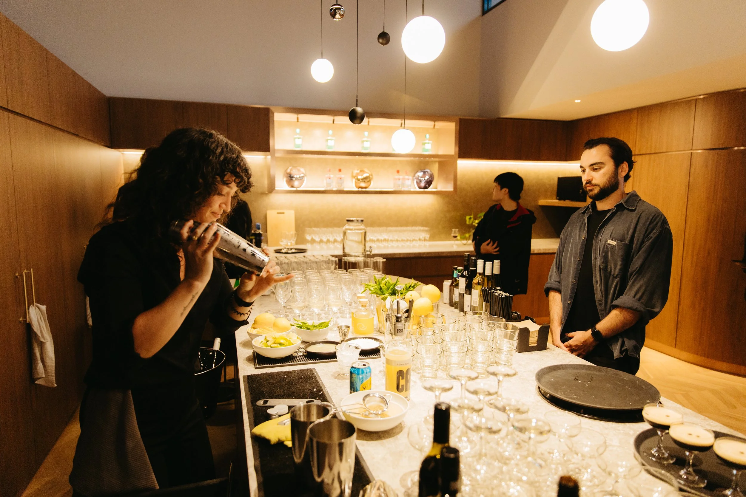Woman pouring a drink at a bar counter with bartending tools and glassware, while two men stand on the other side, in a warmly lit modern bar or restaurant, designed by Raw Process Design, Canberra hospitality interior