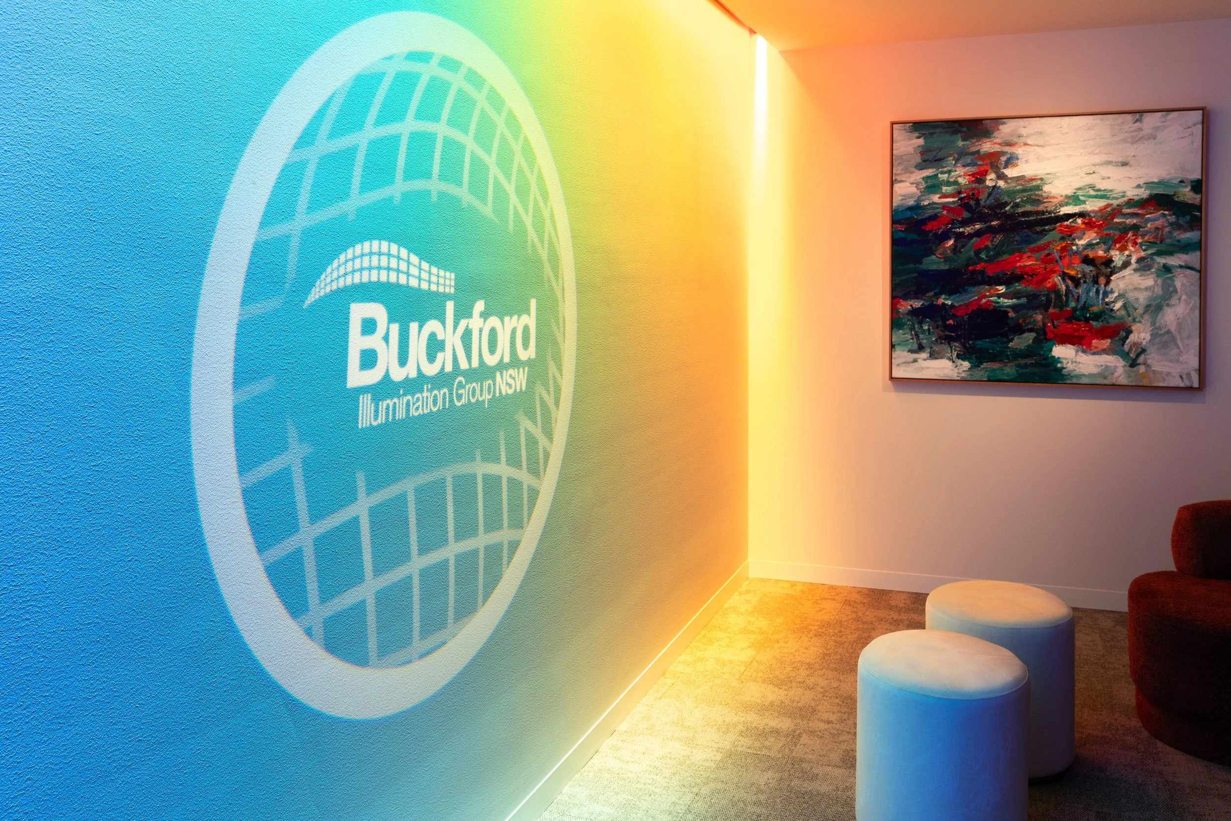 Interior room with wall projection of Buckford Illumination Group NSW logo, colorful abstract painting on wall, two cylindrical seats, and a couch nearby, designed by Raw Process Design, Canberra