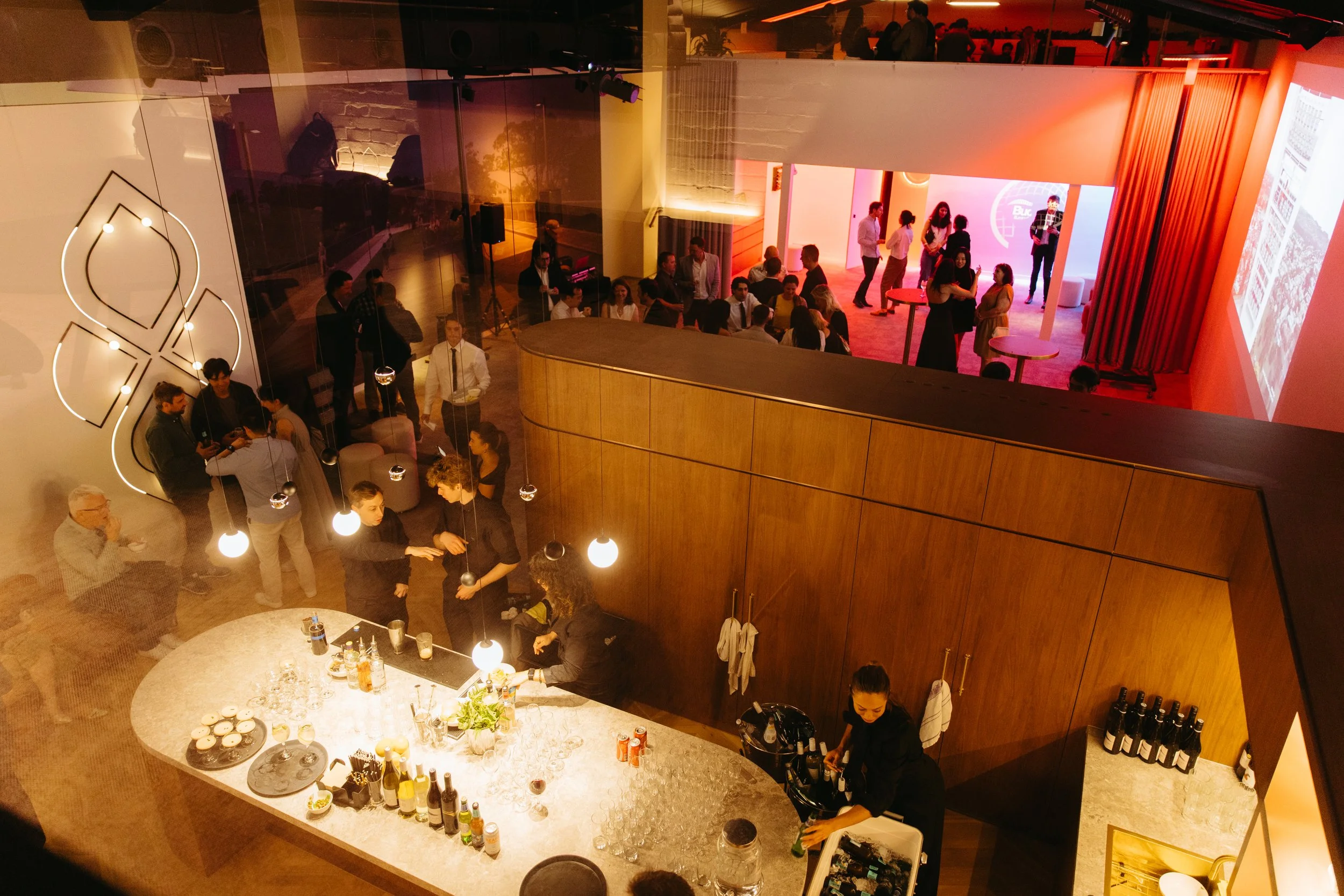 Indoor bar and event space with people socializing, bartenders serving drinks, seating area with guests, large screen projection, and vibrant pink and red lighting, designed by Raw Process Design, Canberra hospitality interiors