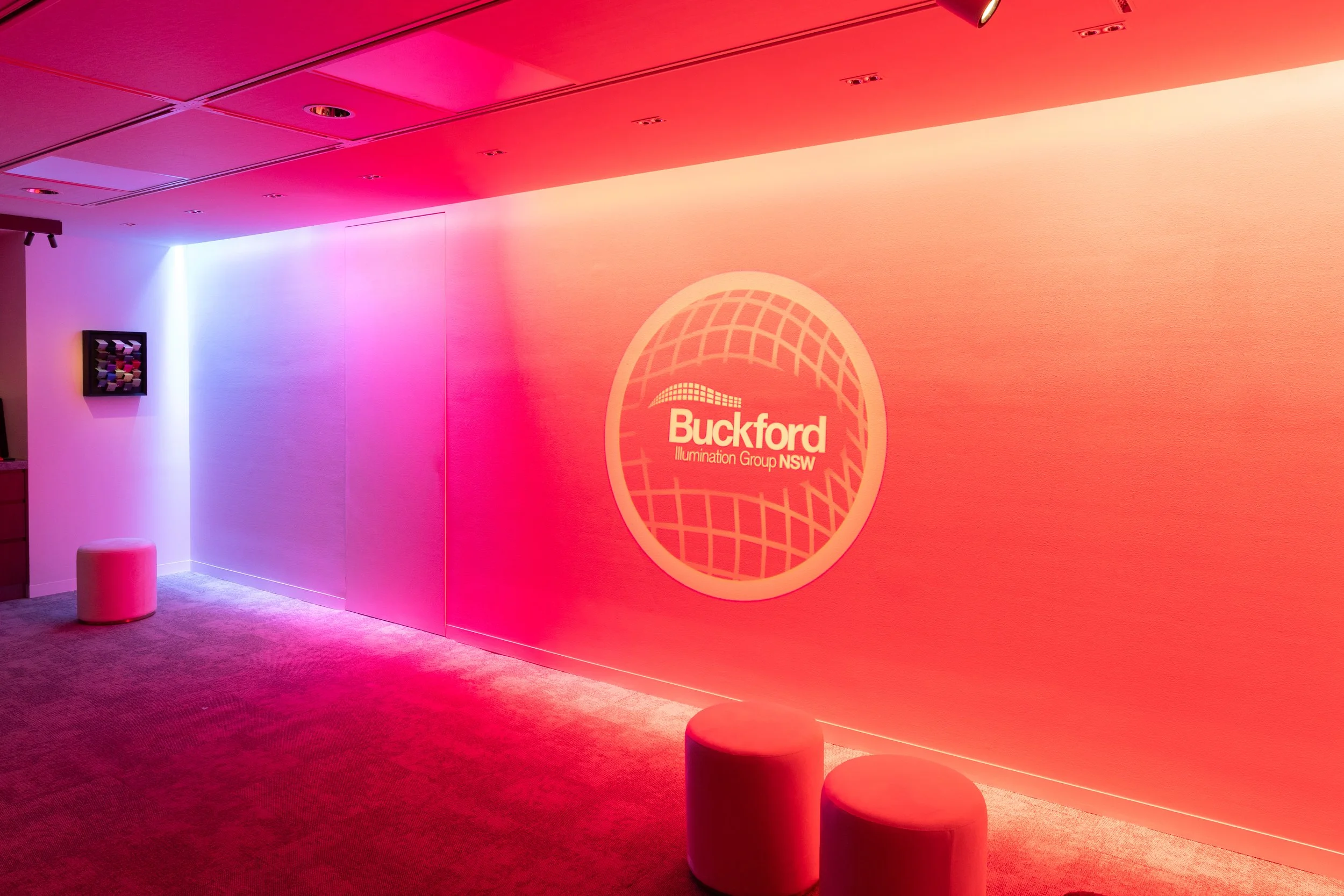 Indoor room with pink and purple lit wall, illuminated Buckford Illumination Group NSW logo, two round stools on carpeted floor, and small artwork on side wall, designed by Raw Process Design, Canberra interiors