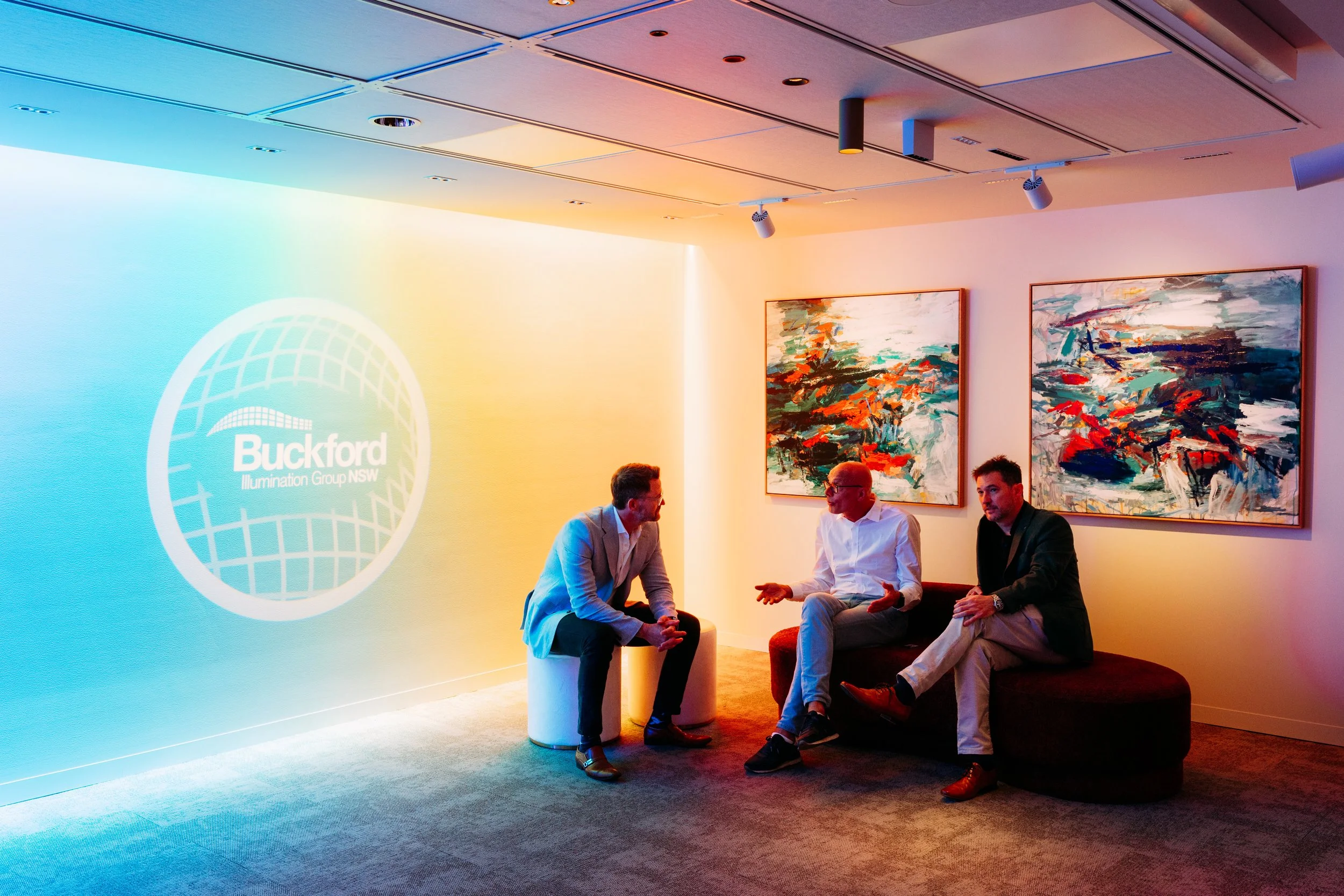 Three men engaged in conversation in an art gallery, with colorful abstract paintings on the wall and a projection on the left reading 'Buckford Illumination Group NSW'.
