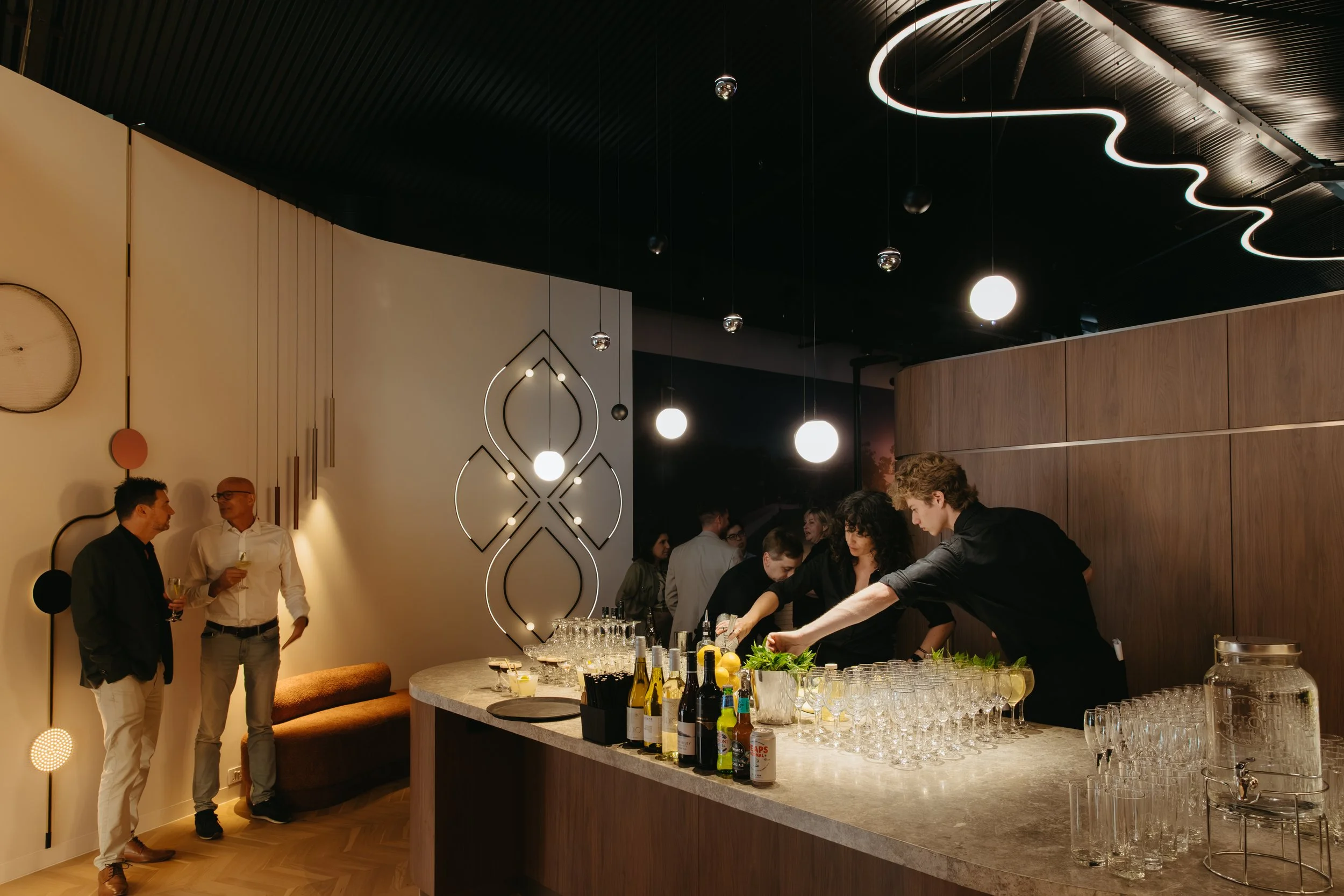 Indoor bar with people socializing, bartenders preparing drinks, modern lighting, and wooden wall decor, designed by Raw Process Design, Canberra hospitality interior