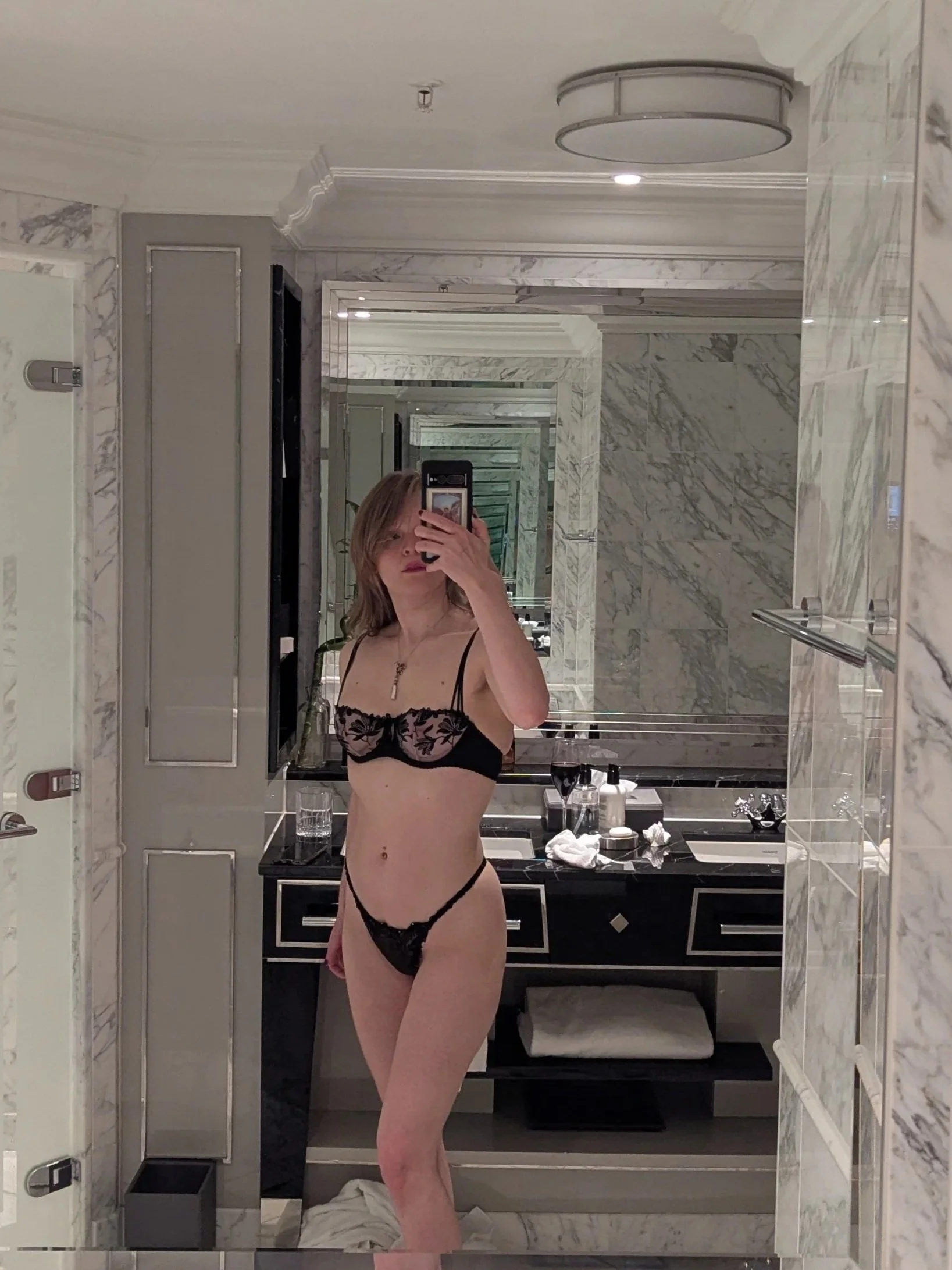 Mirror selfie of Lyra Bergman, hairy escort, wearing lingerie, taken in a marble bathroom