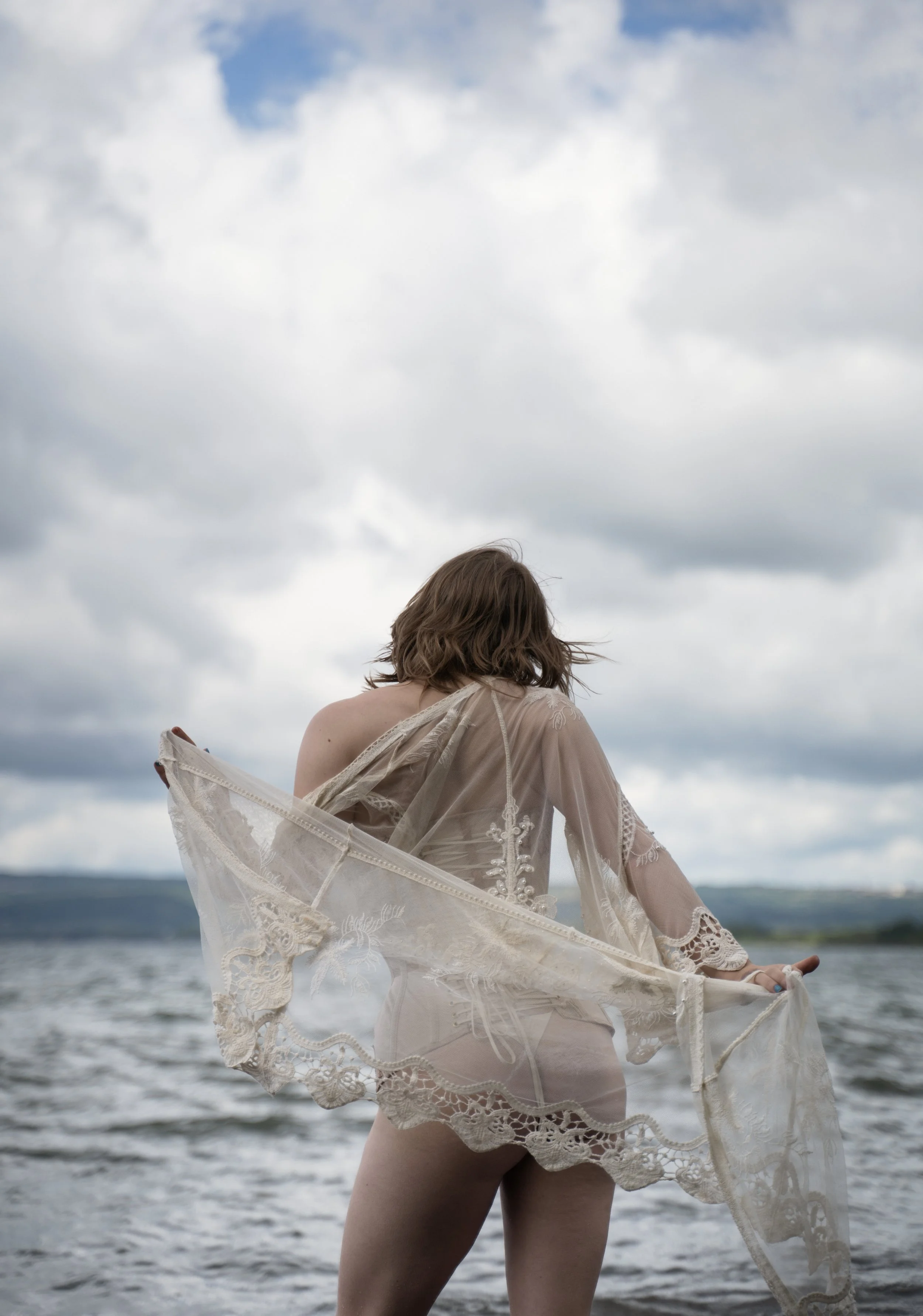 Lyra Bergman, independent Berlin escort, stands in the water of a lake, her back facing the viewer, dressed in lace and a thonf