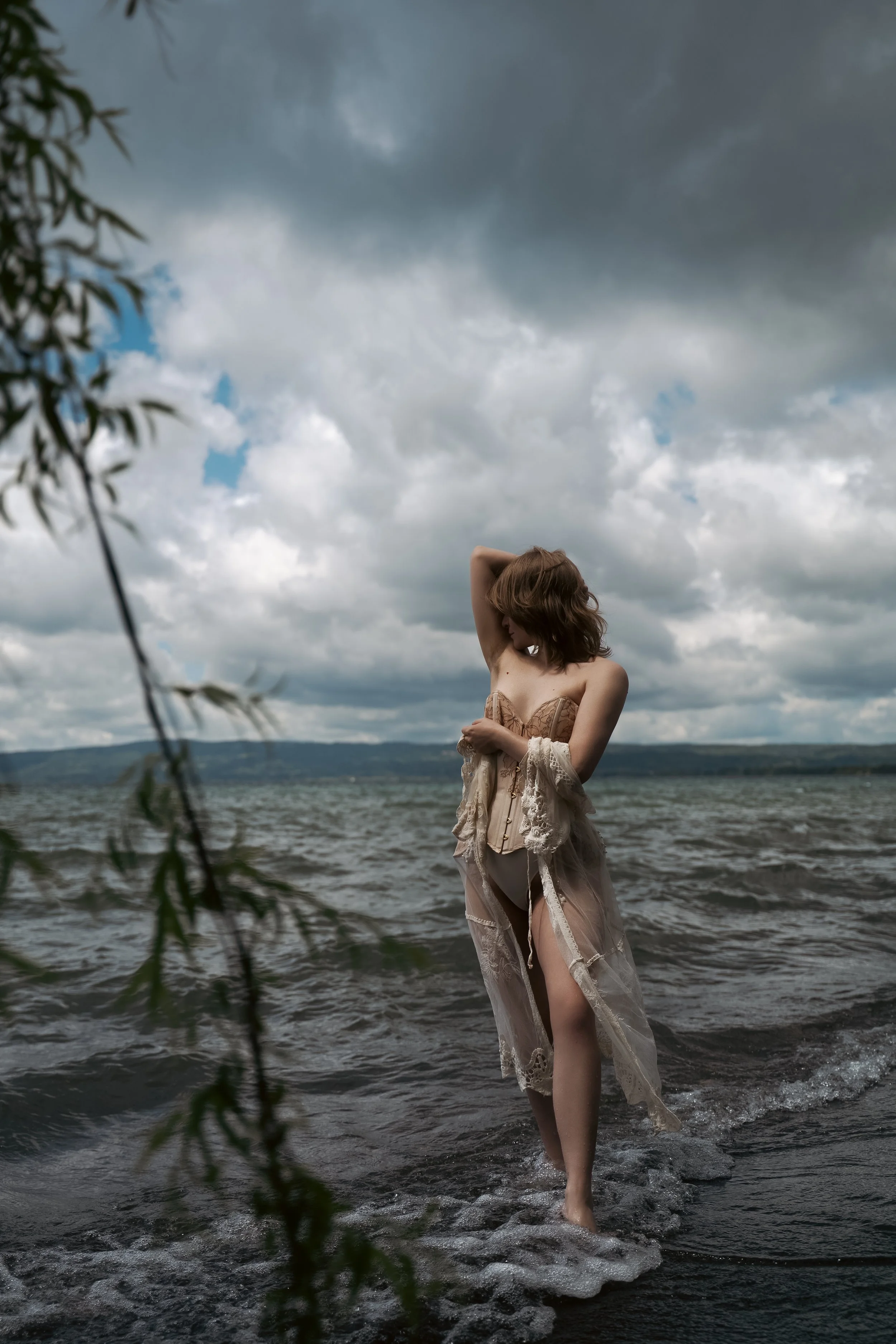 Luxury GFE escort Lyra Bergman walks on the edge of the water in a lake in Italy, wearing white underwear and a white corset, draped in a sheer vintage lingerie jacket