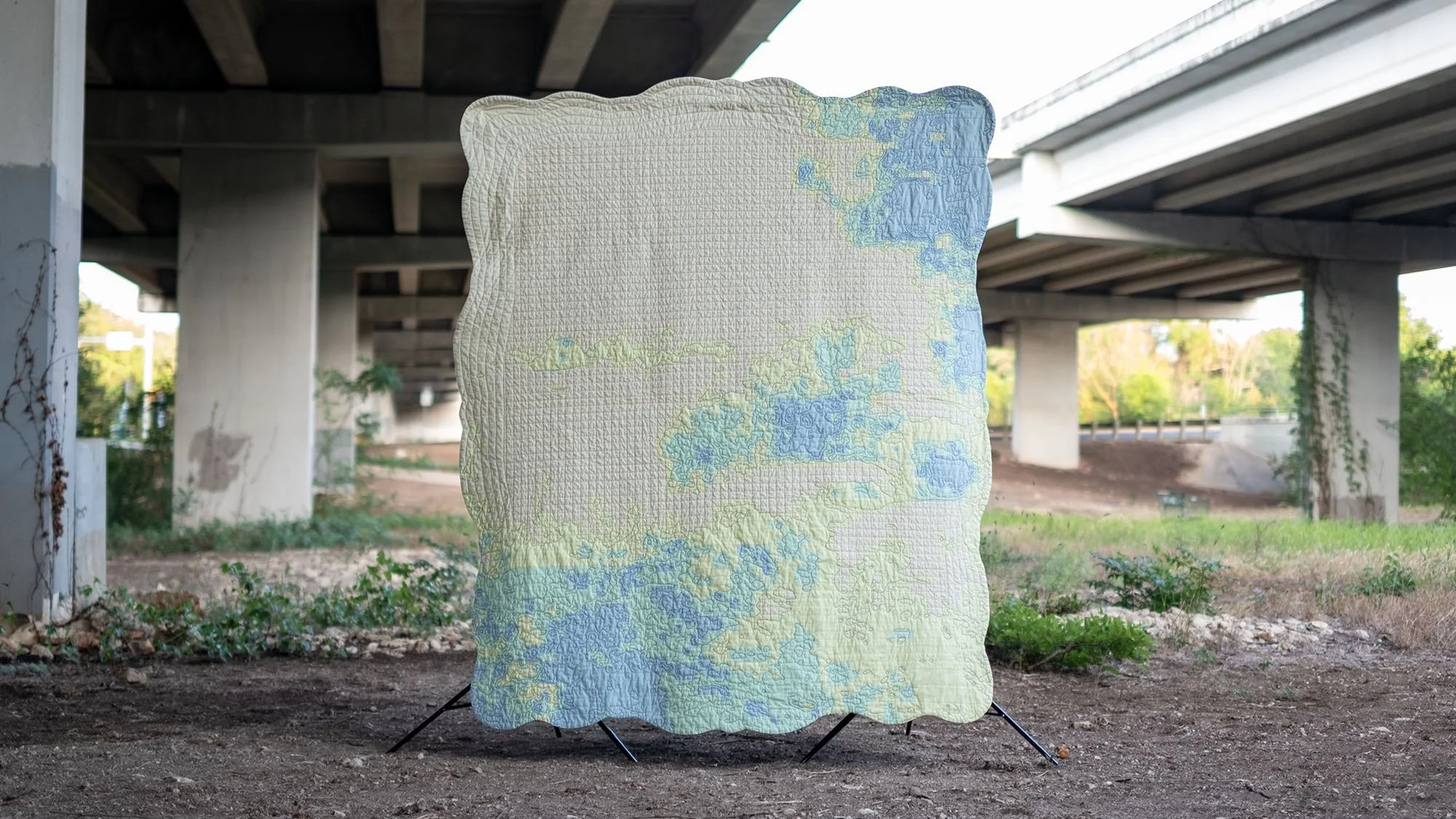 Front of the quilt suspended beneath an overpass, showing the curved frame border surrounding pale blue and green pieced forms.