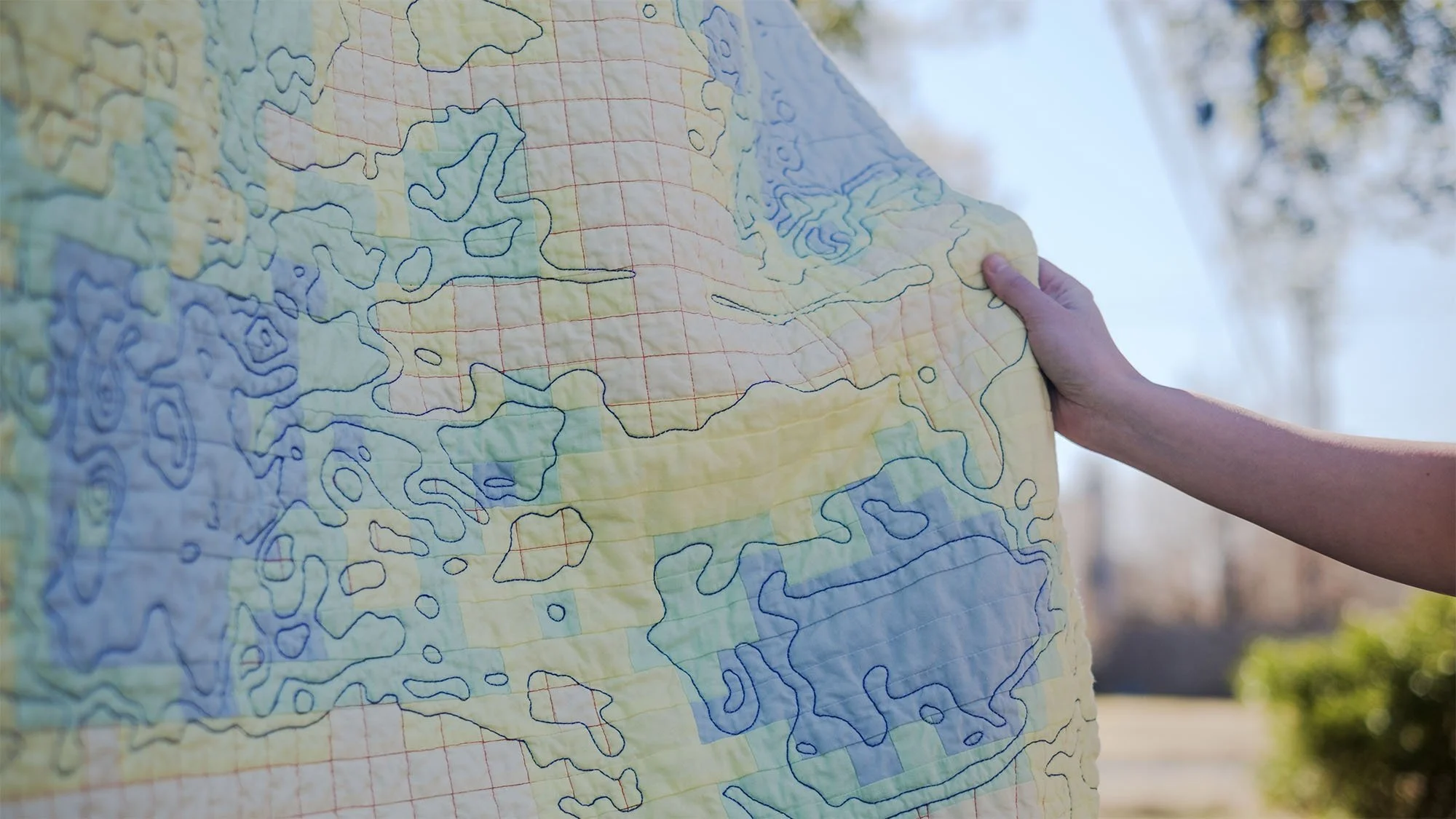 Detail of the quilt draped in sunlight showing red grid quilting layered beneath blue contour quilting.